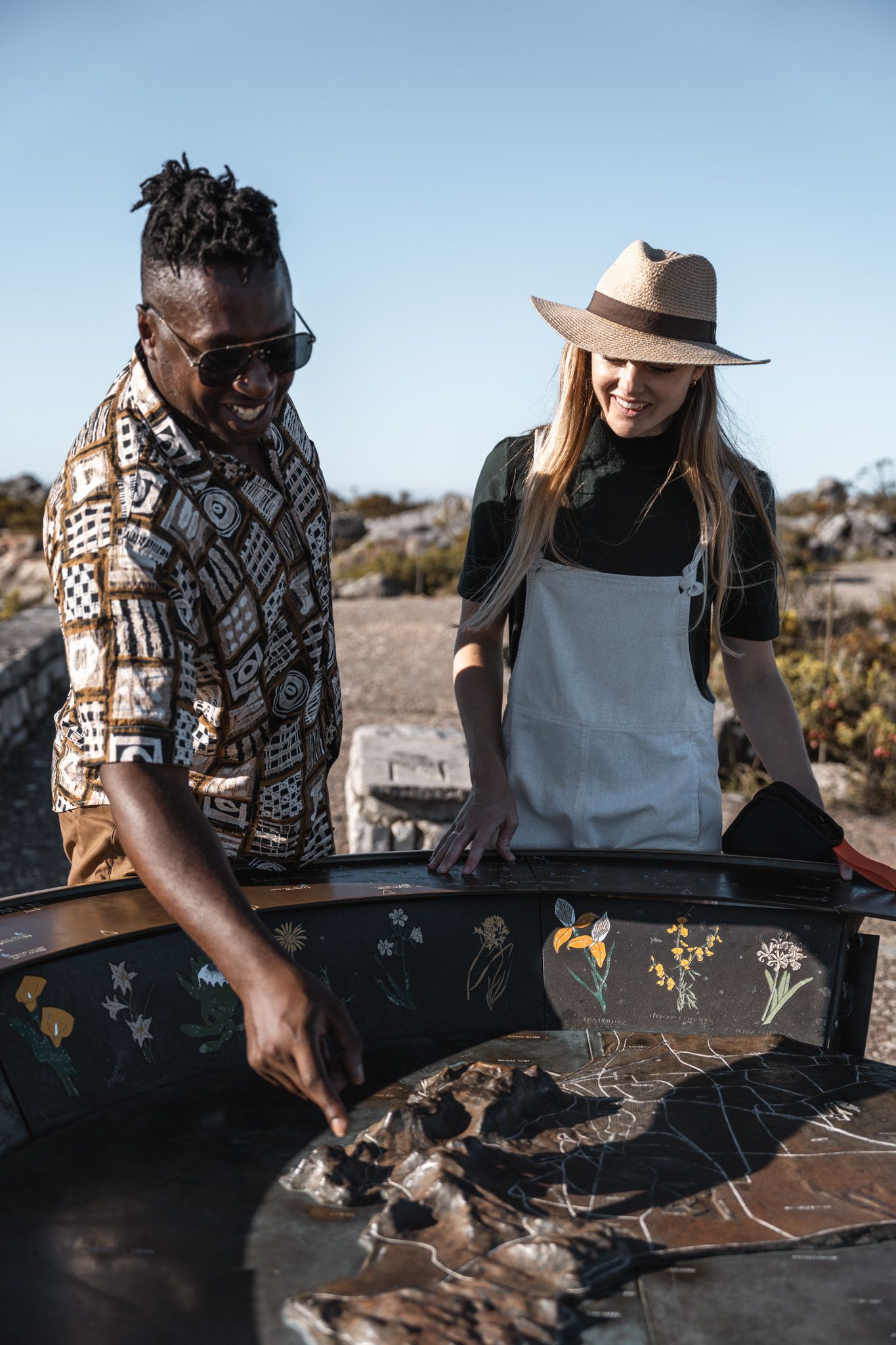 Two people prepare food at a decorated outdoor table in a desert setting during a sunny day.