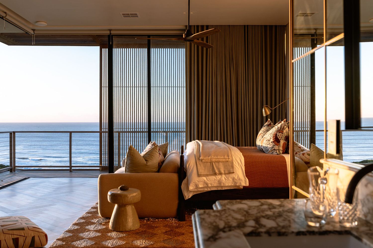 Modern coastal bedroom with panoramic ocean views, beige sectional sofa, and warm natural lighting.