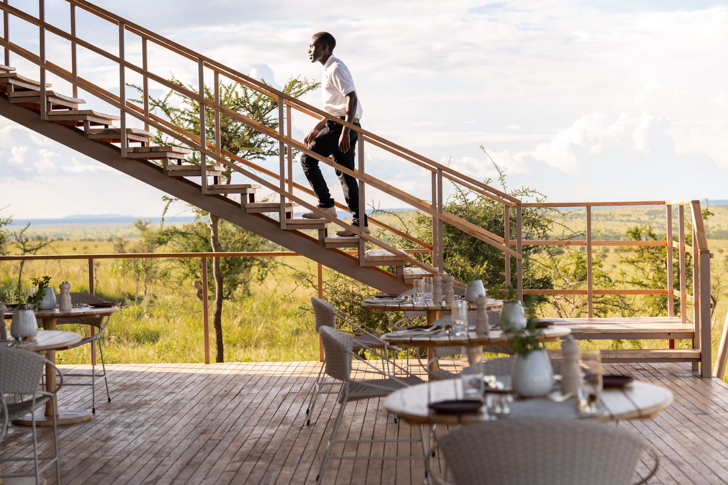Staff member walks up wooden staircase at safari lodge with scenic savanna views.