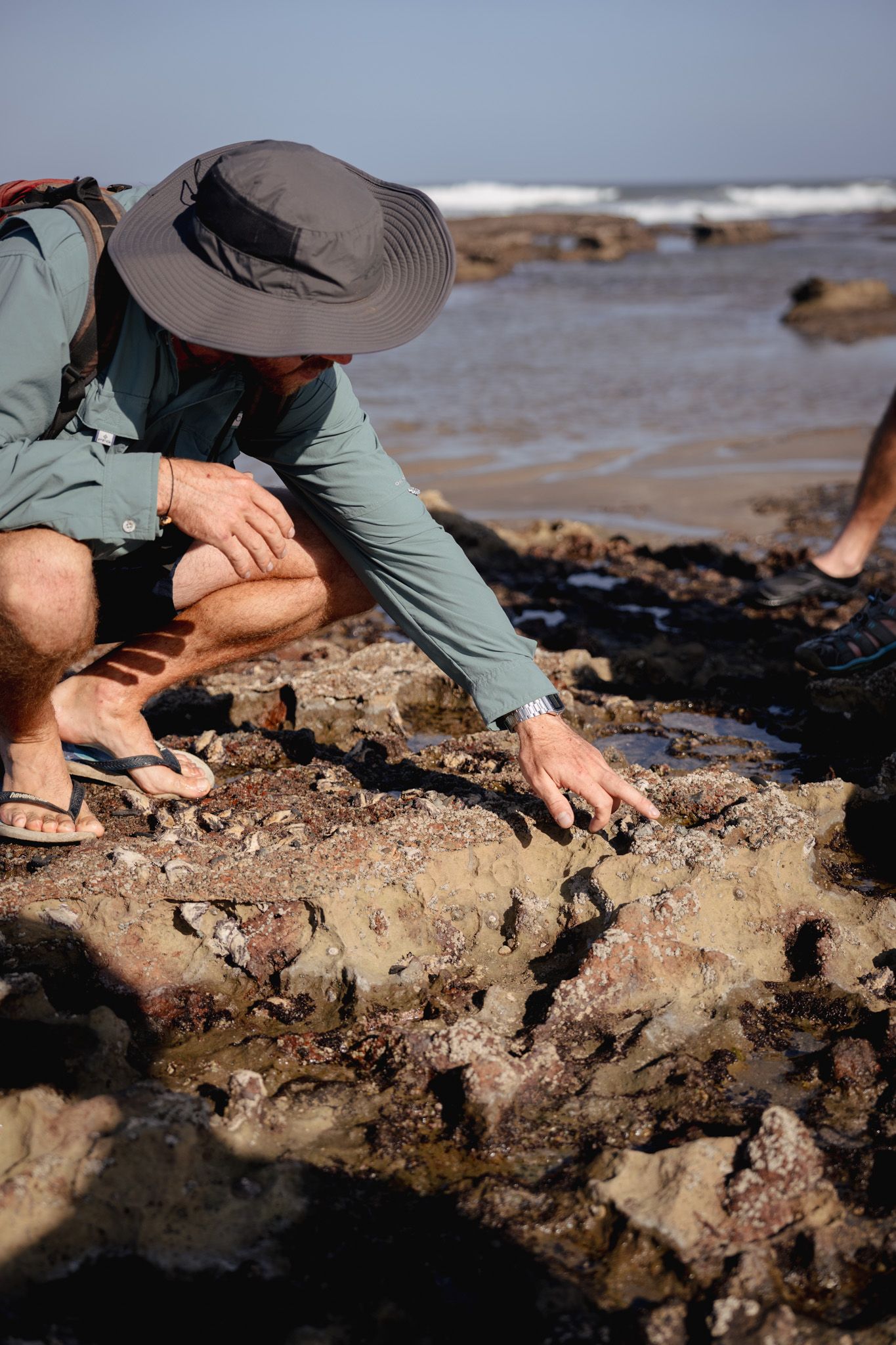 Person in sun hat examining rocky coastal formations at low tide.