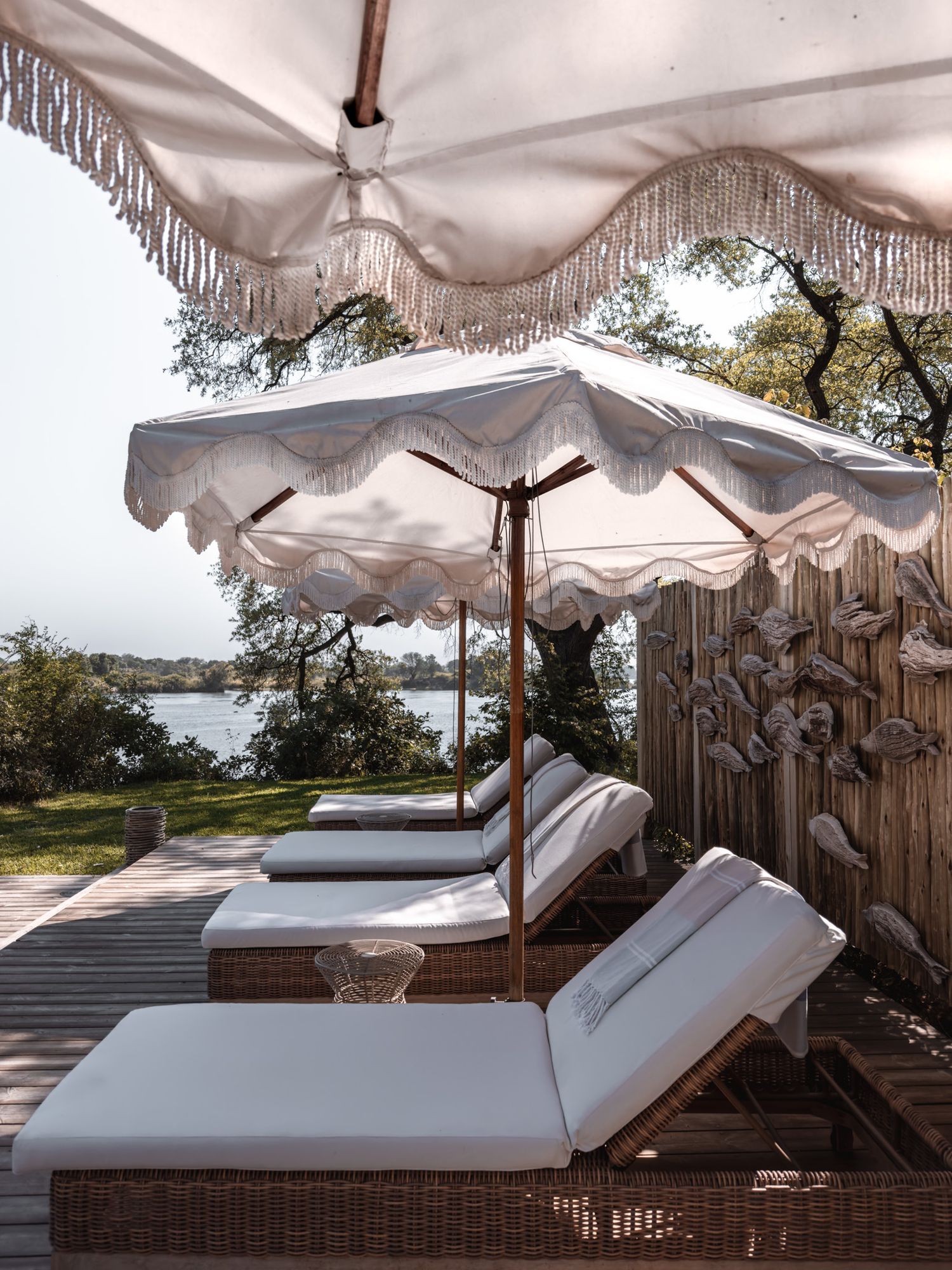 Luxurious poolside lounge chairs with white parasols and decorative fringe overlooking a waterfront.