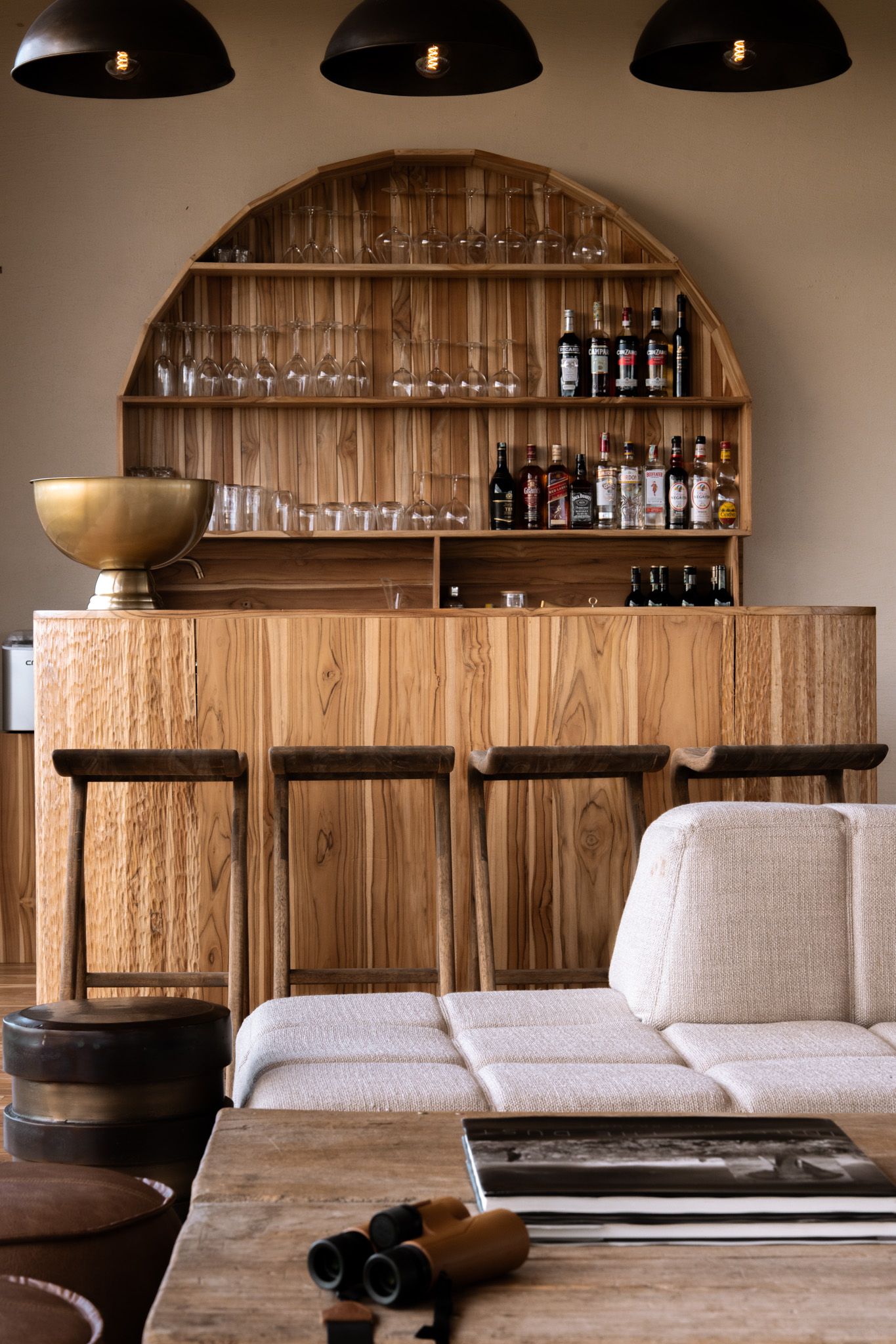 Modern wooden home bar with arched shelving displaying glasses and bottles under black pendant lights.