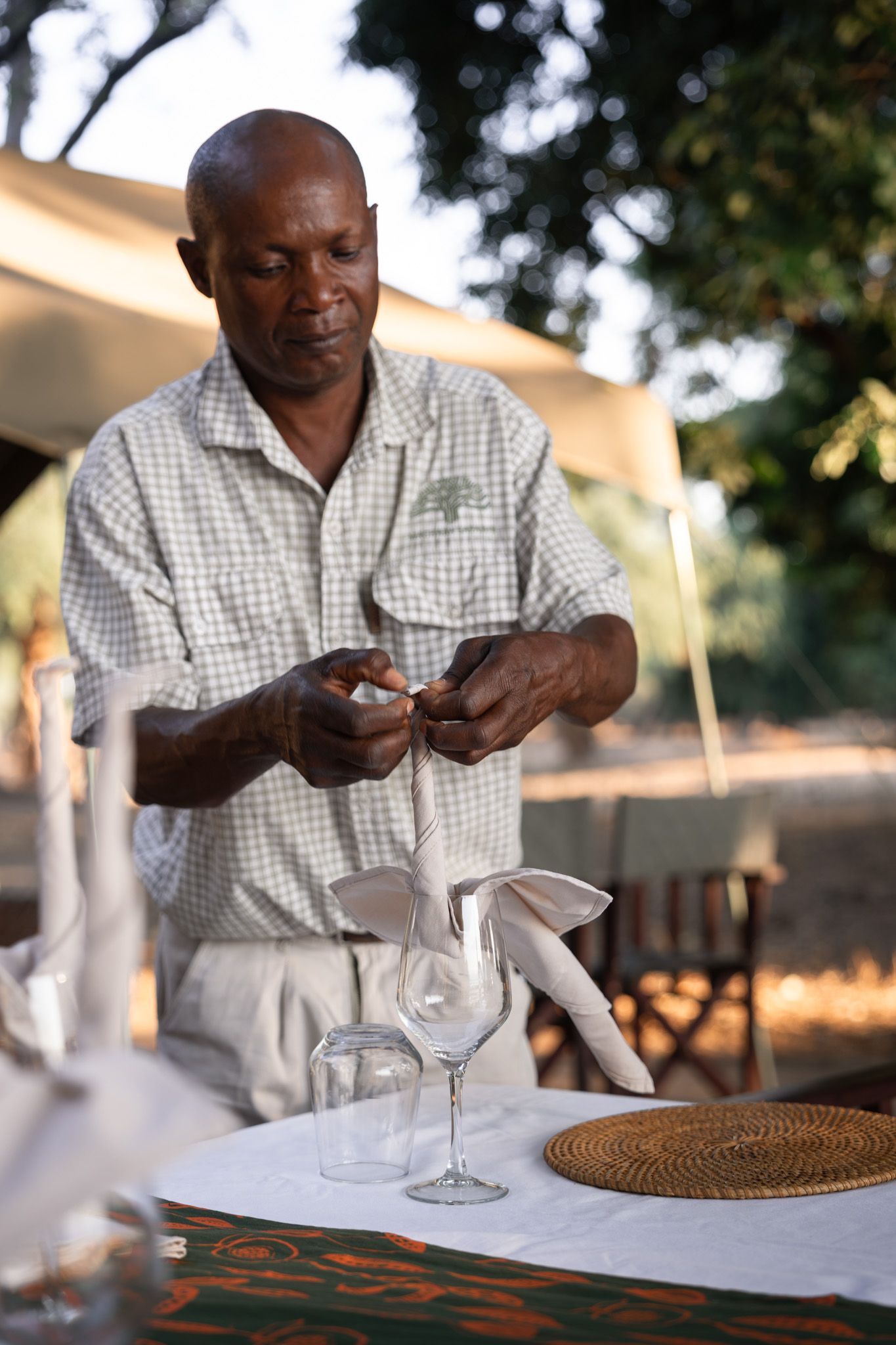 Staff member sets formal dining table with wine glasses at outdoor safari venue.