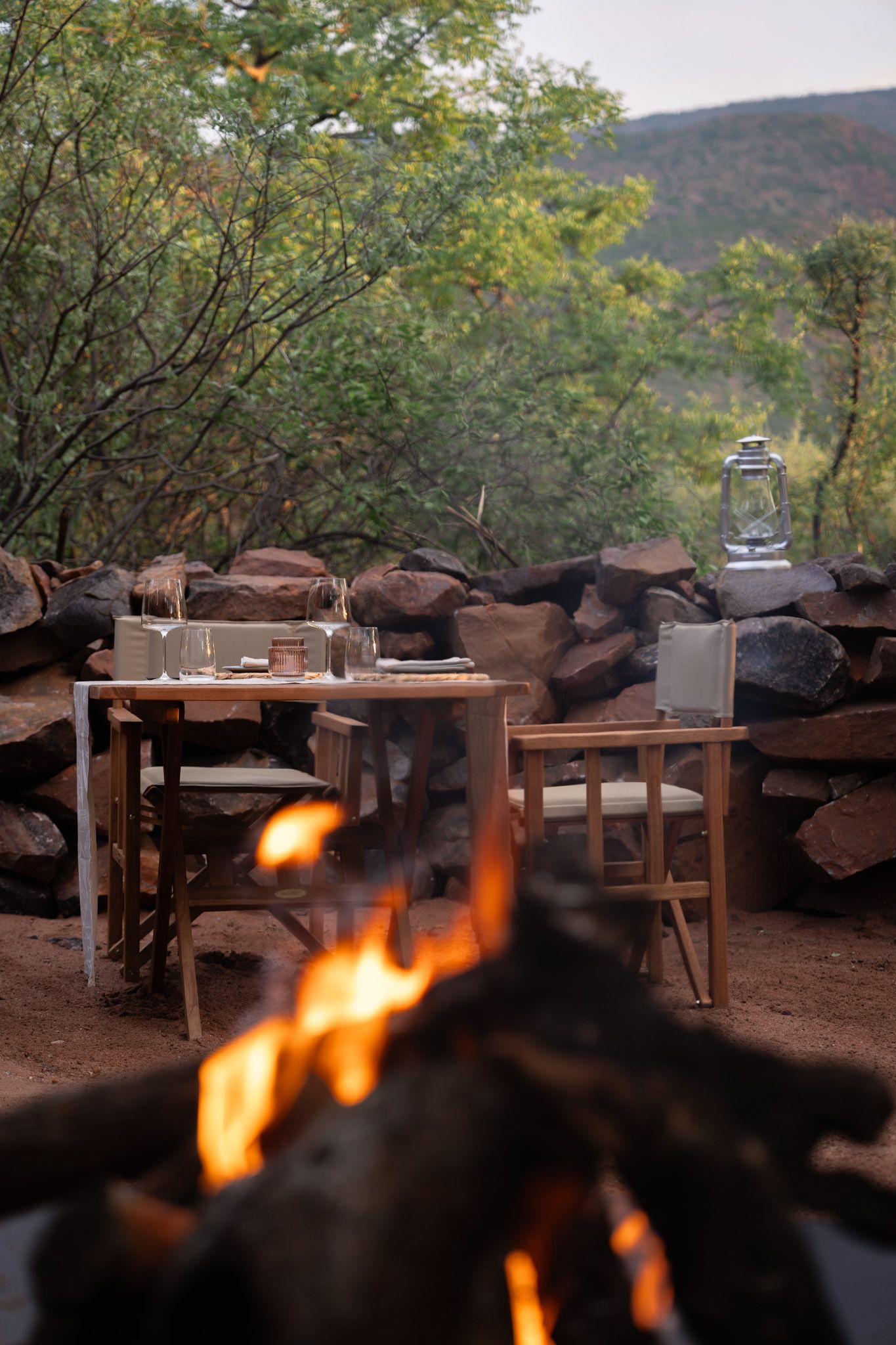 Outdoor dining table set near campfire with mountain views at dusk.
