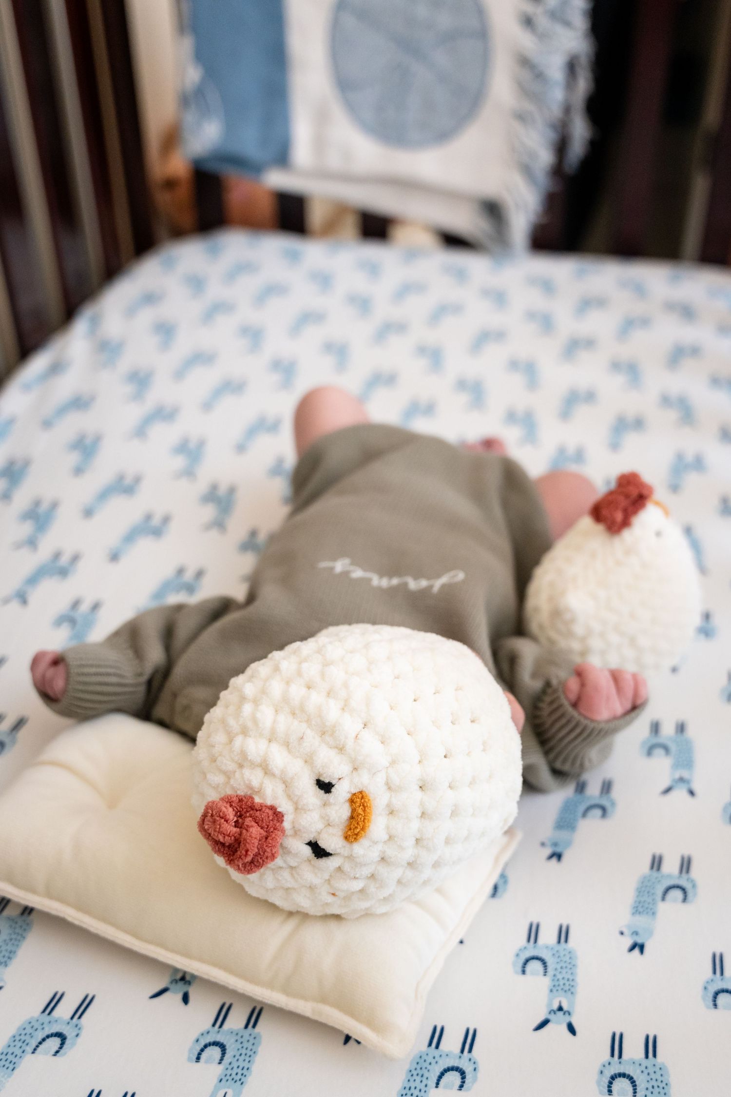 Baby wearing gray outfit laying on blue patterned bedding next to white crocheted snowman plush.
