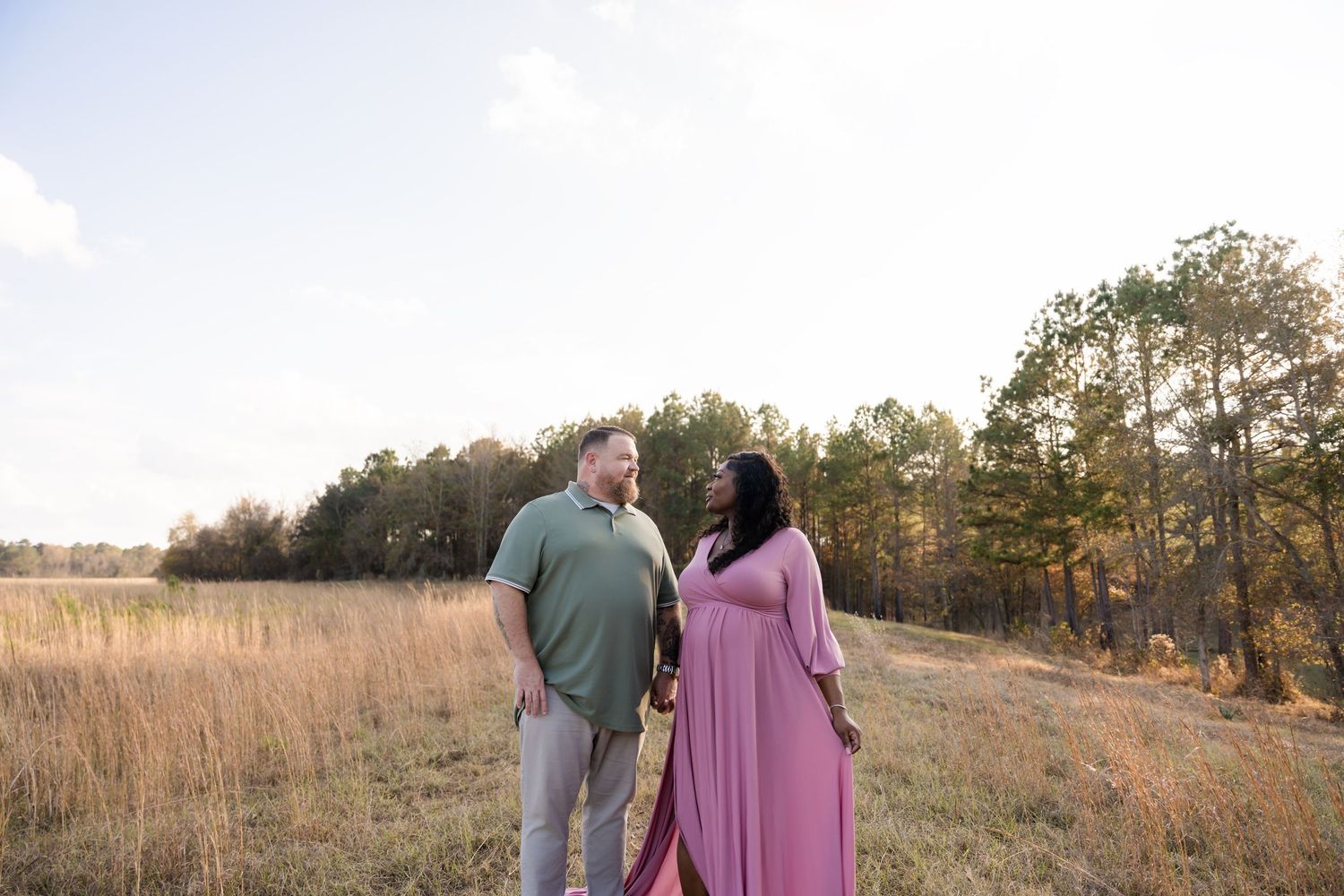 Couple standing in golden field at sunset, woman in pink maxi dress, man in green polo, trees in background.