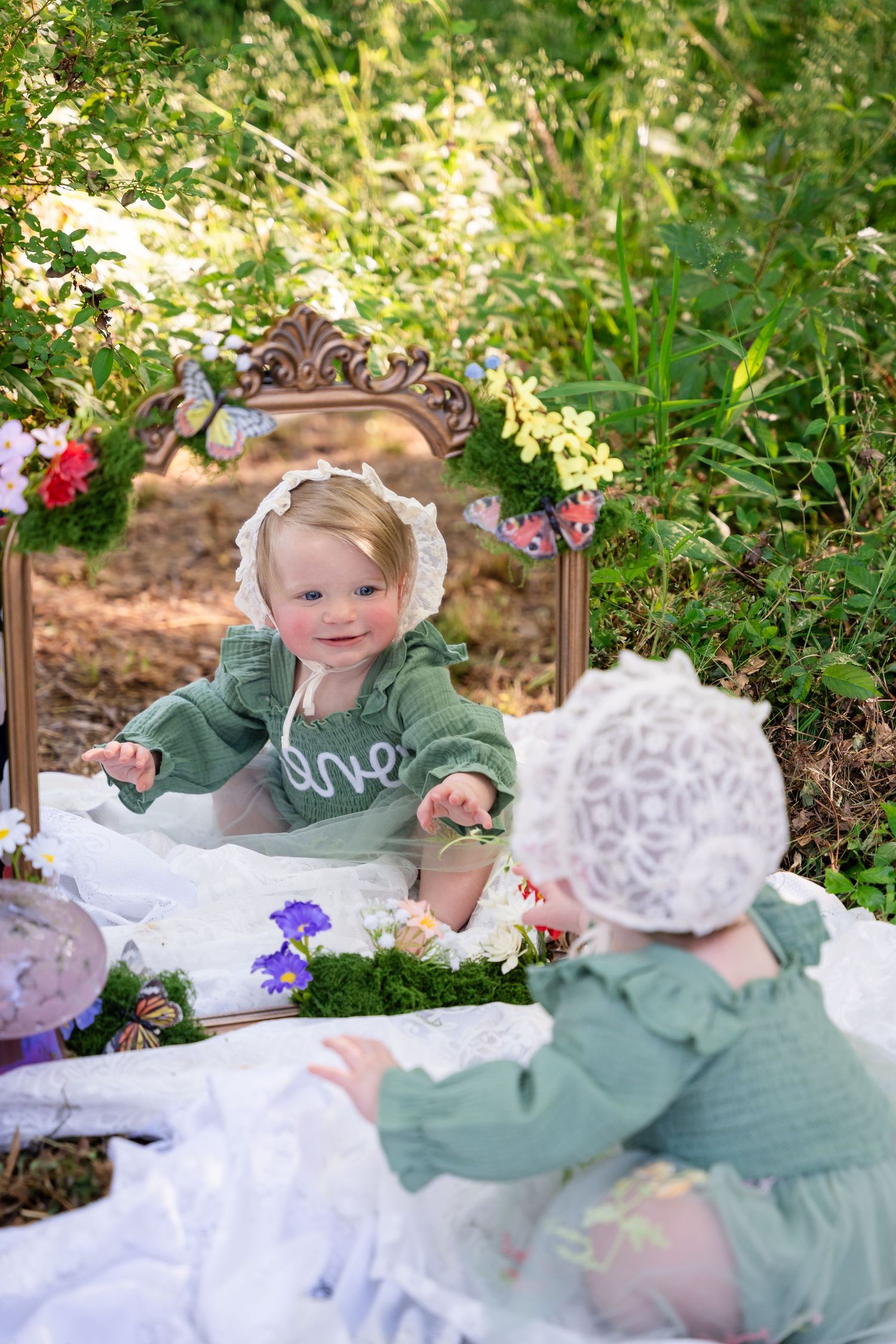 Smiling baby girl in green outfit and white bonnet reaches toward ornate mirror outdoors in a whimsical garden scene.