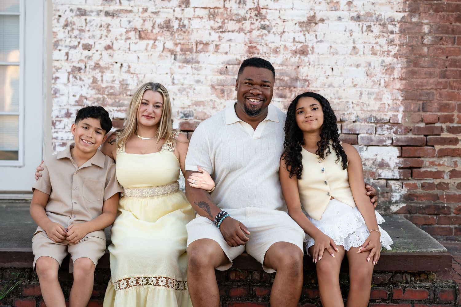 Family of four smiling together on brick steps, wearing coordinated cream and white outfits for a portrait photo.