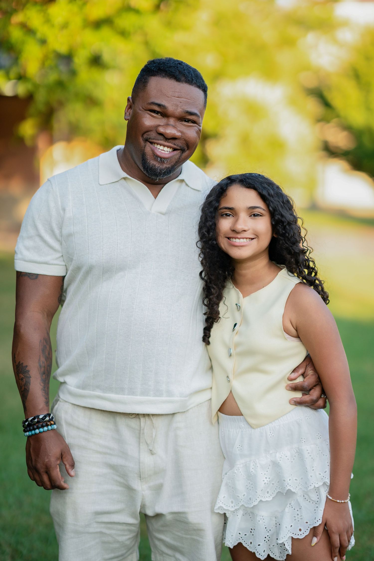 A smiling man in white outfit poses with a curly-haired girl in white attire in a sunny park.