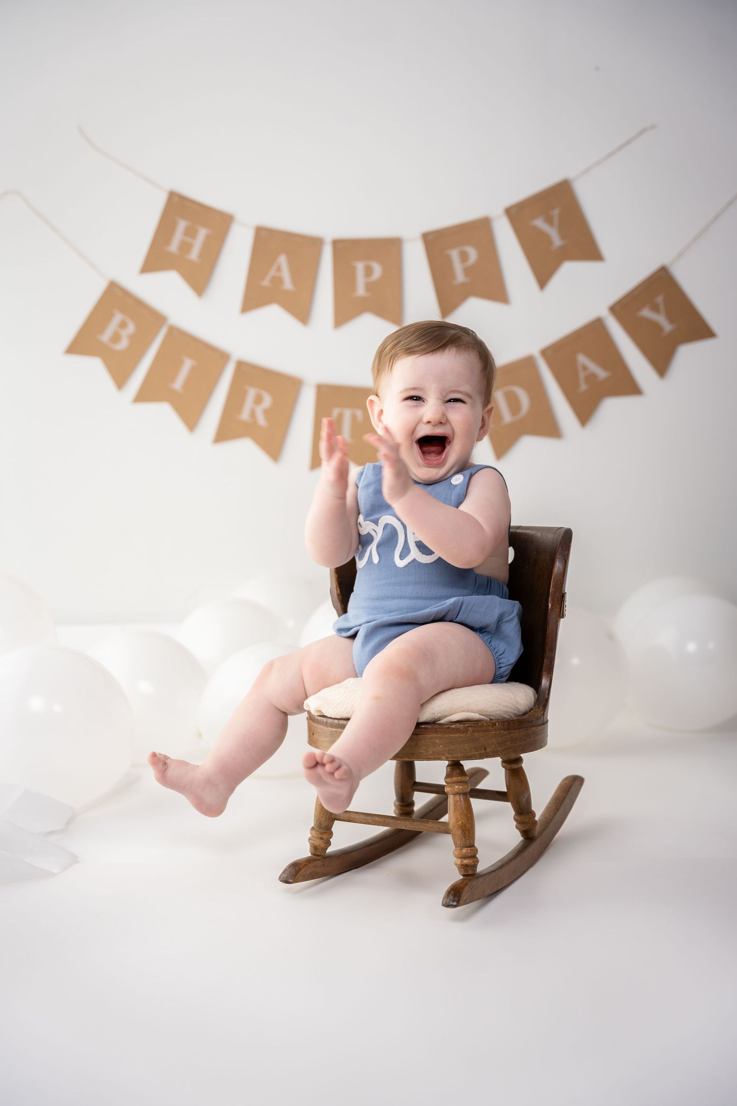 Birthday celebration setup with kraft paper banner, white balloons, and small wooden rocking chair against white backdrop.