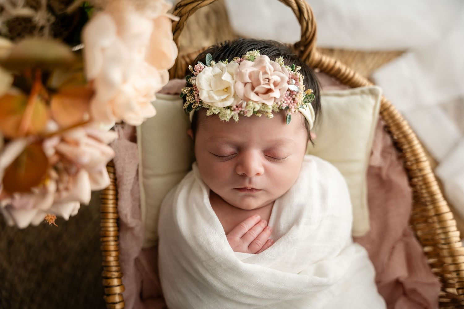 Newborn baby sleeping peacefully in wicker basket surrounded by soft pink flowers and white fabric.