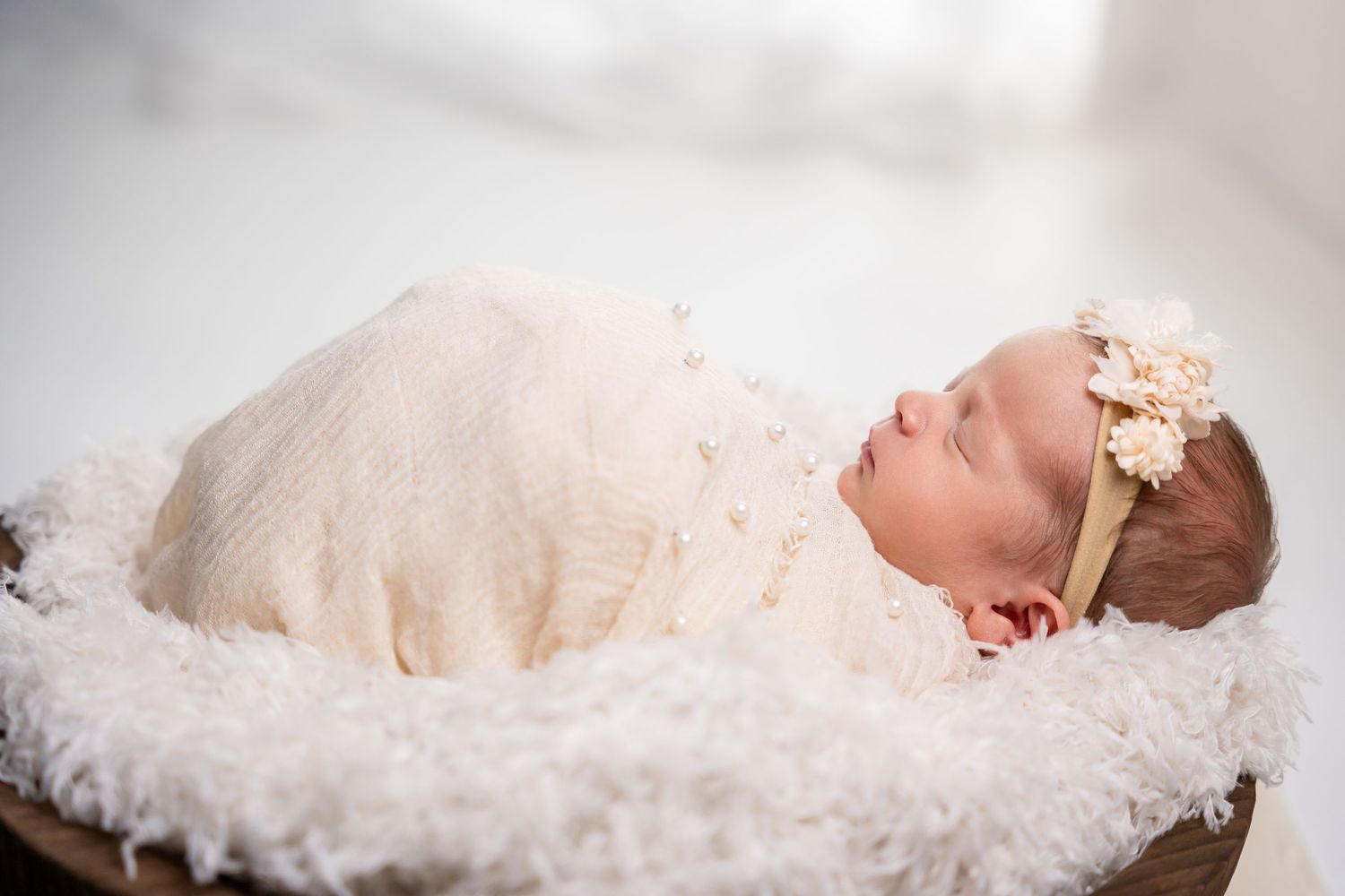 Sleeping newborn wrapped in cream lace overlay on fluffy white surface.