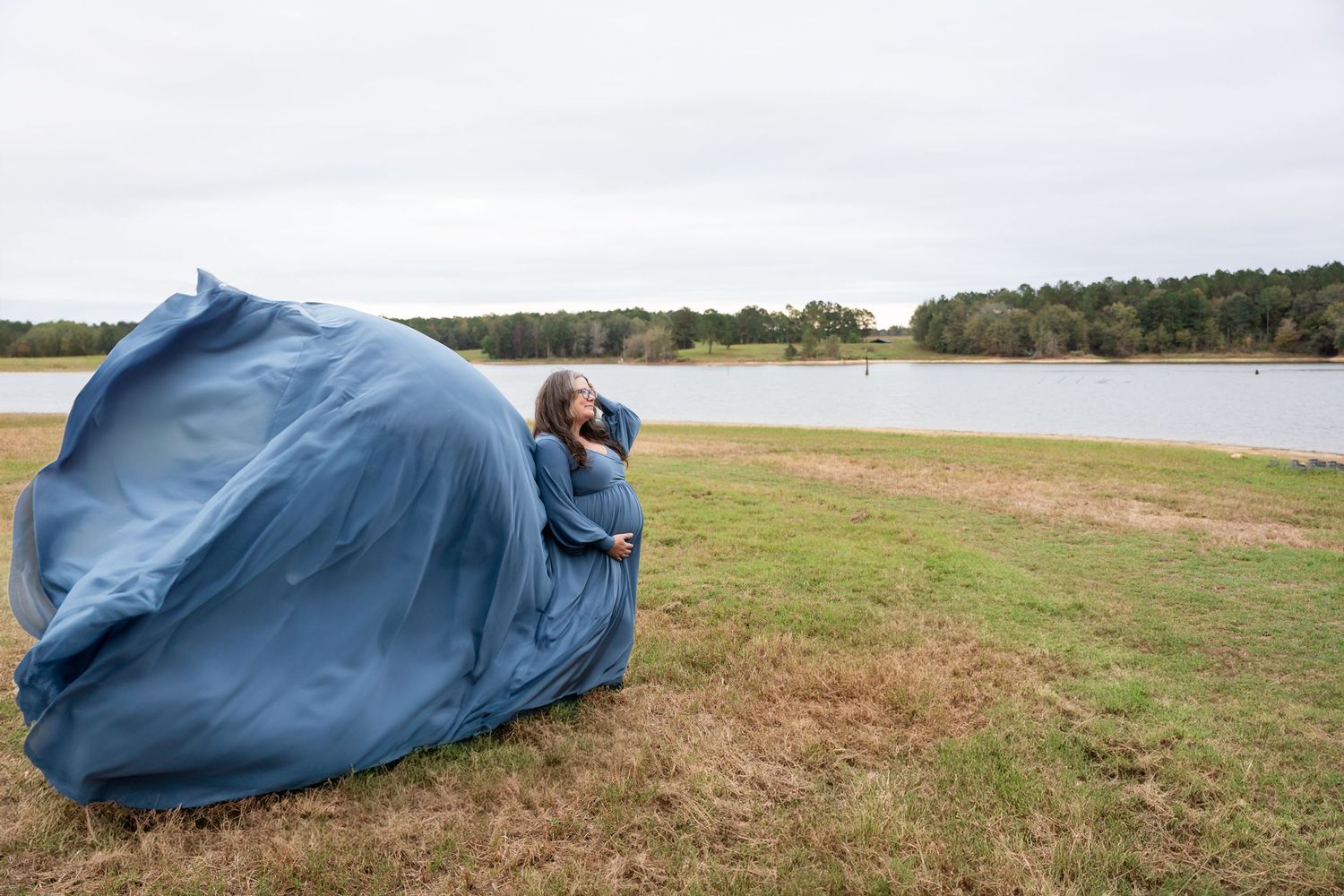 Blue dress billows dramatically against scenic lake and grass landscape.