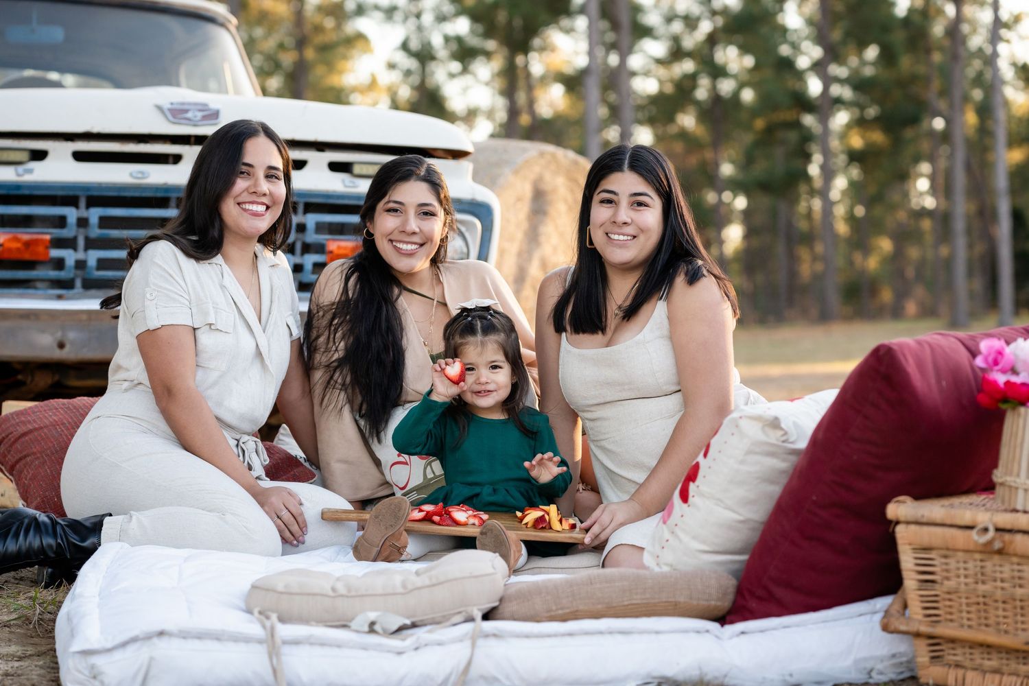 Group seated on white blankets having outdoor picnic near vintage pickup truck.