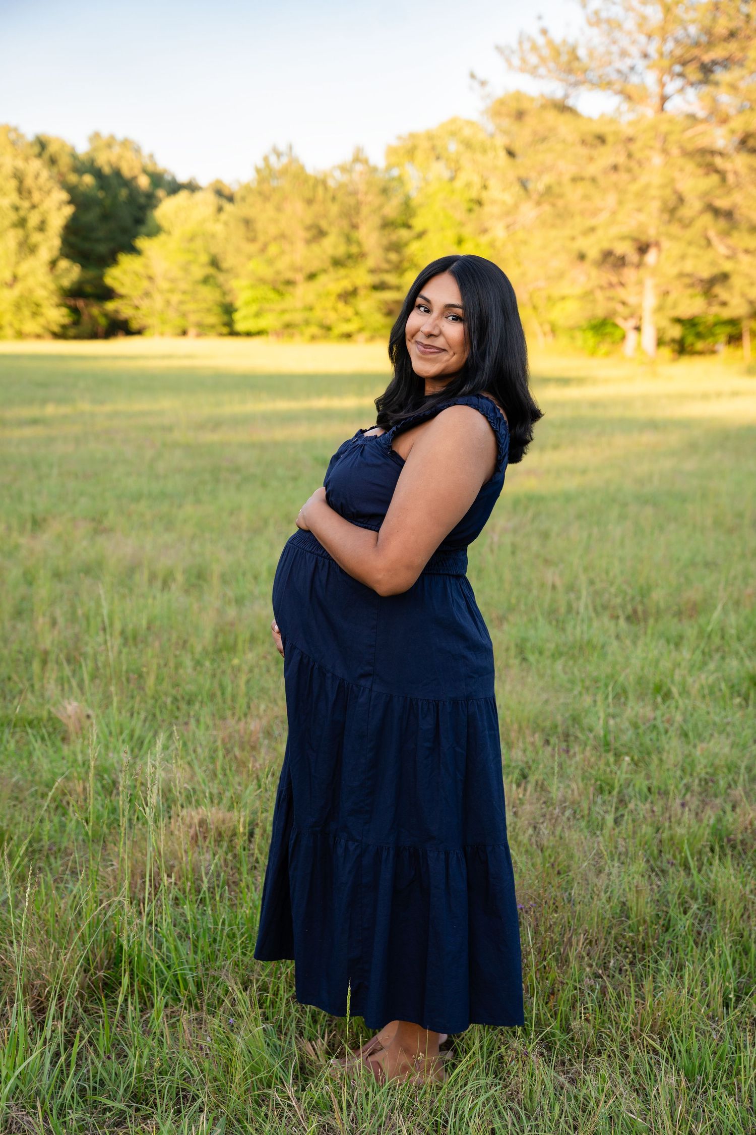 Smiling pregnant woman in navy blue dress cradling baby bump in lush green field at golden hour.