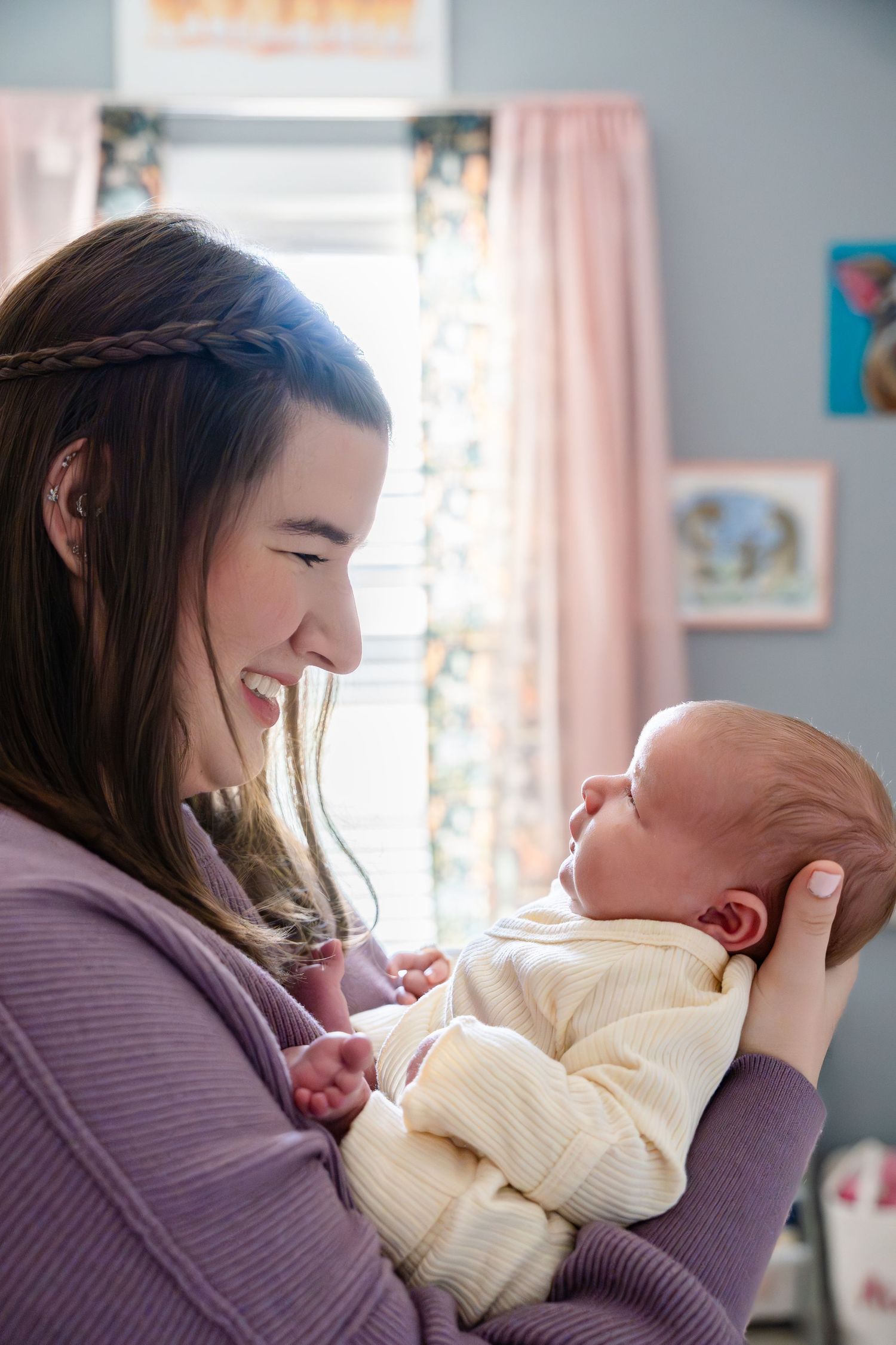 Smiling young mother holding her newborn baby in a cozy nursery with pink curtains and soft lighting.