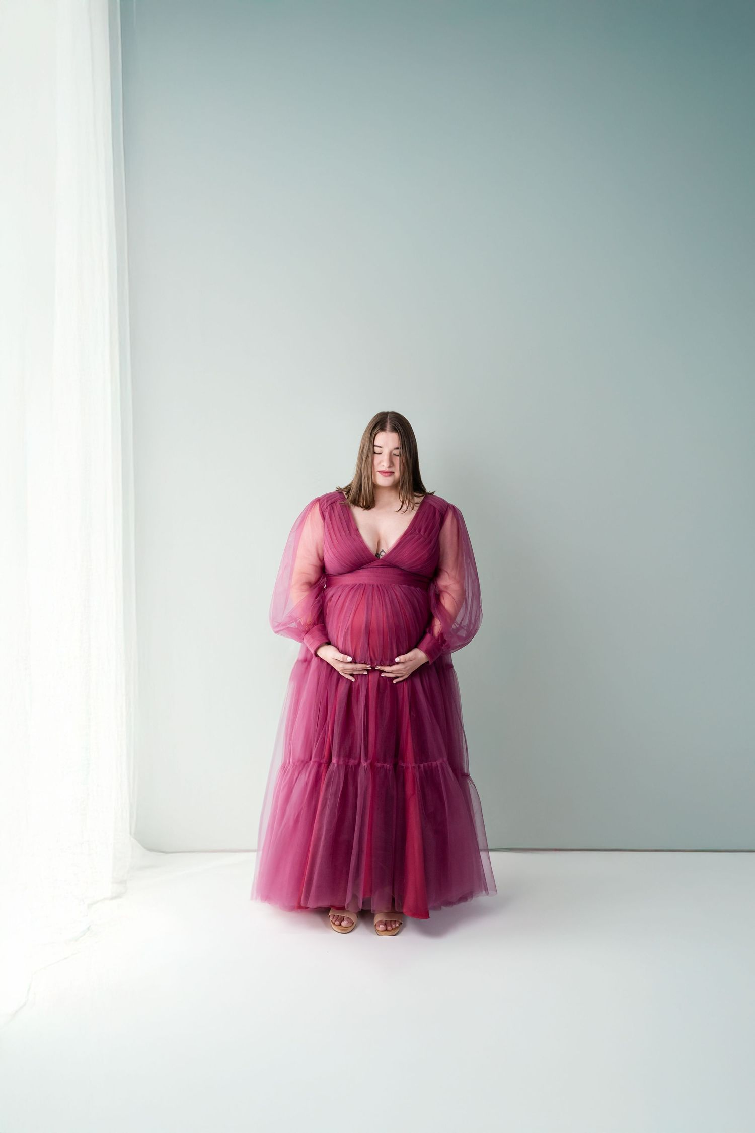 A woman in a flowing burgundy tulle gown poses gracefully in a minimalist white studio setting.