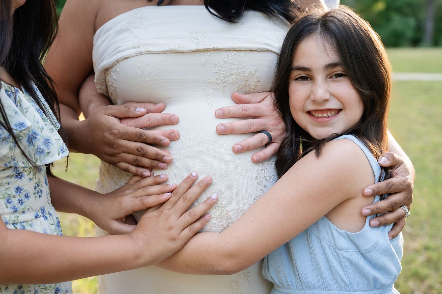 Smiling young girl in a blue dress hugs a pregnant woman's belly outdoors during a sunny maternity photo session with family.
