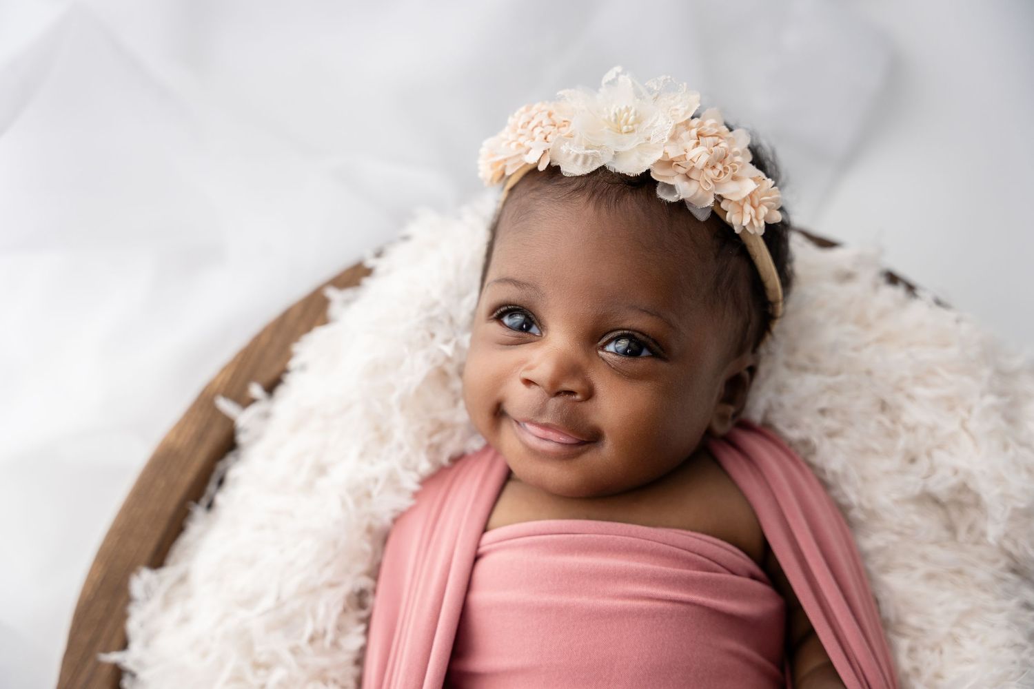 Baby wearing pink wrap and floral headband smiles while lying in wooden bowl with white fluffy blanket.