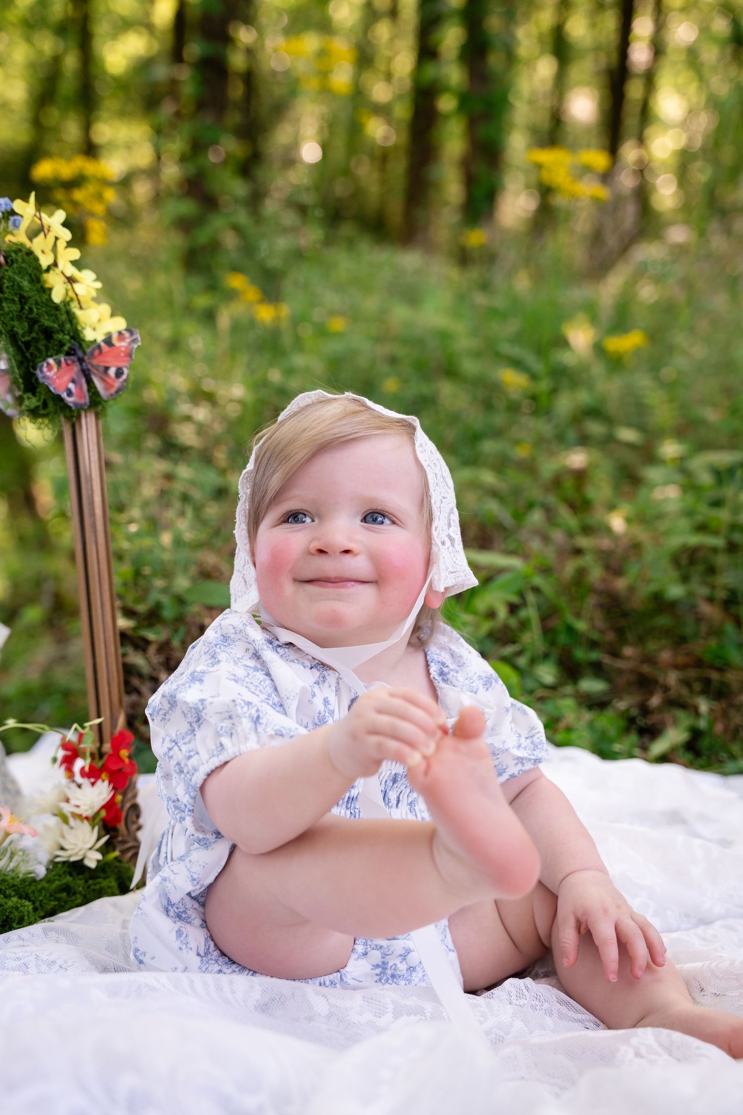 Adorable baby in a white lace bonnet and floral dress sitting outdoors on a white blanket in a lush green forest.