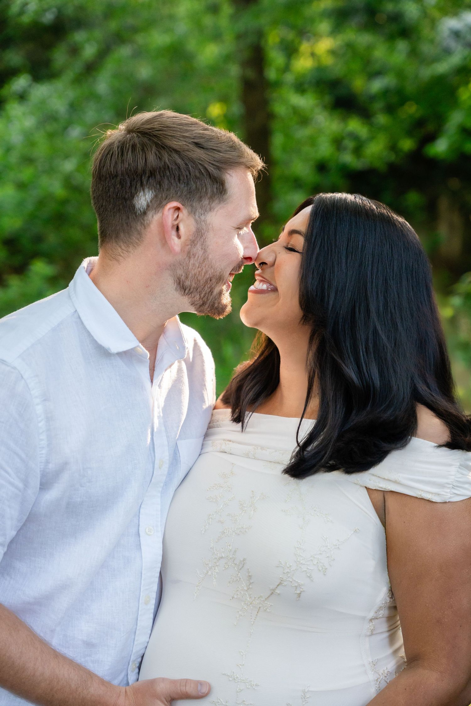 A couple in white outfits share a tender nose-to-nose moment outdoors, smiling with lush green trees in the background.