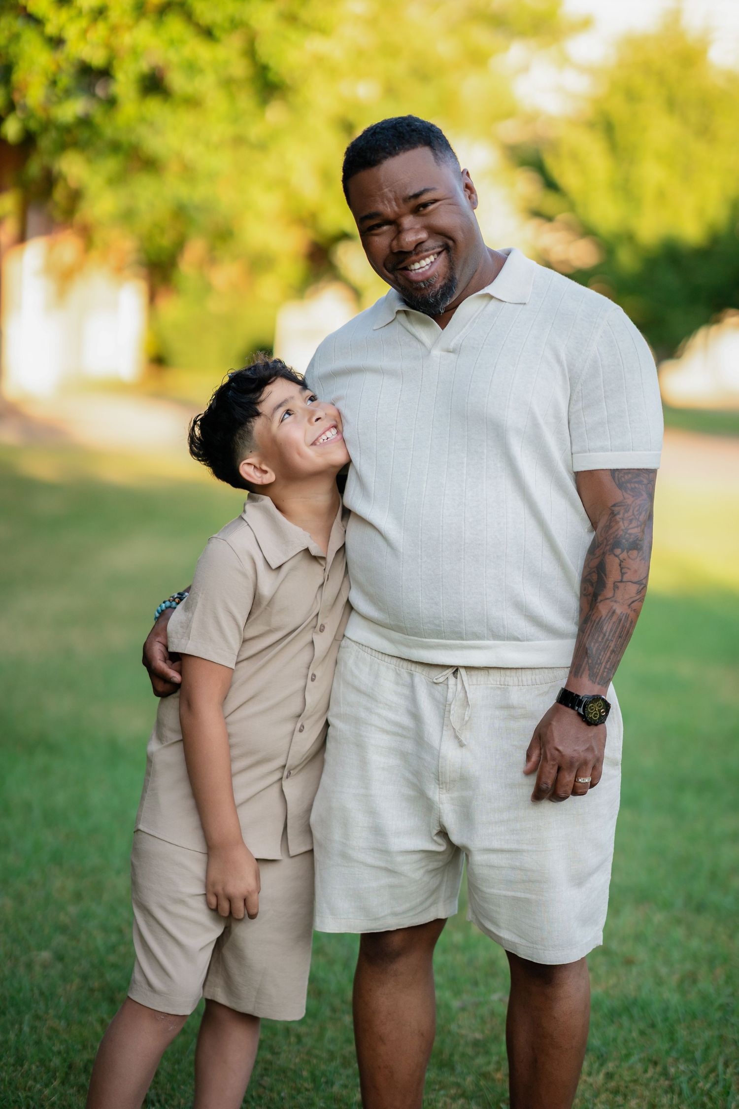 A smiling man and boy in matching beige outfits share a warm embrace outdoors on green grass.