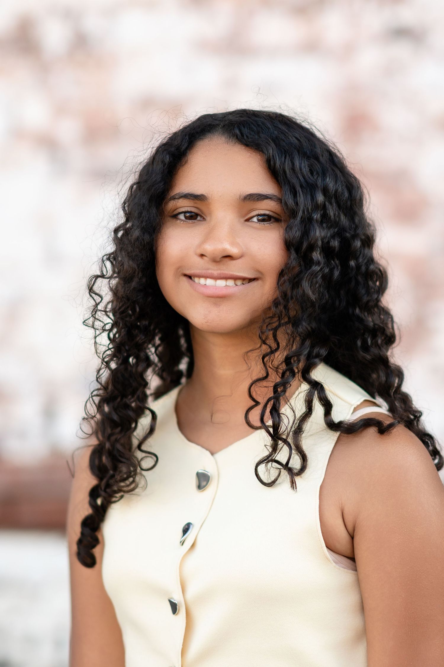 Young woman with long curly black hair smiling, wearing a cream sleeveless top outdoors.