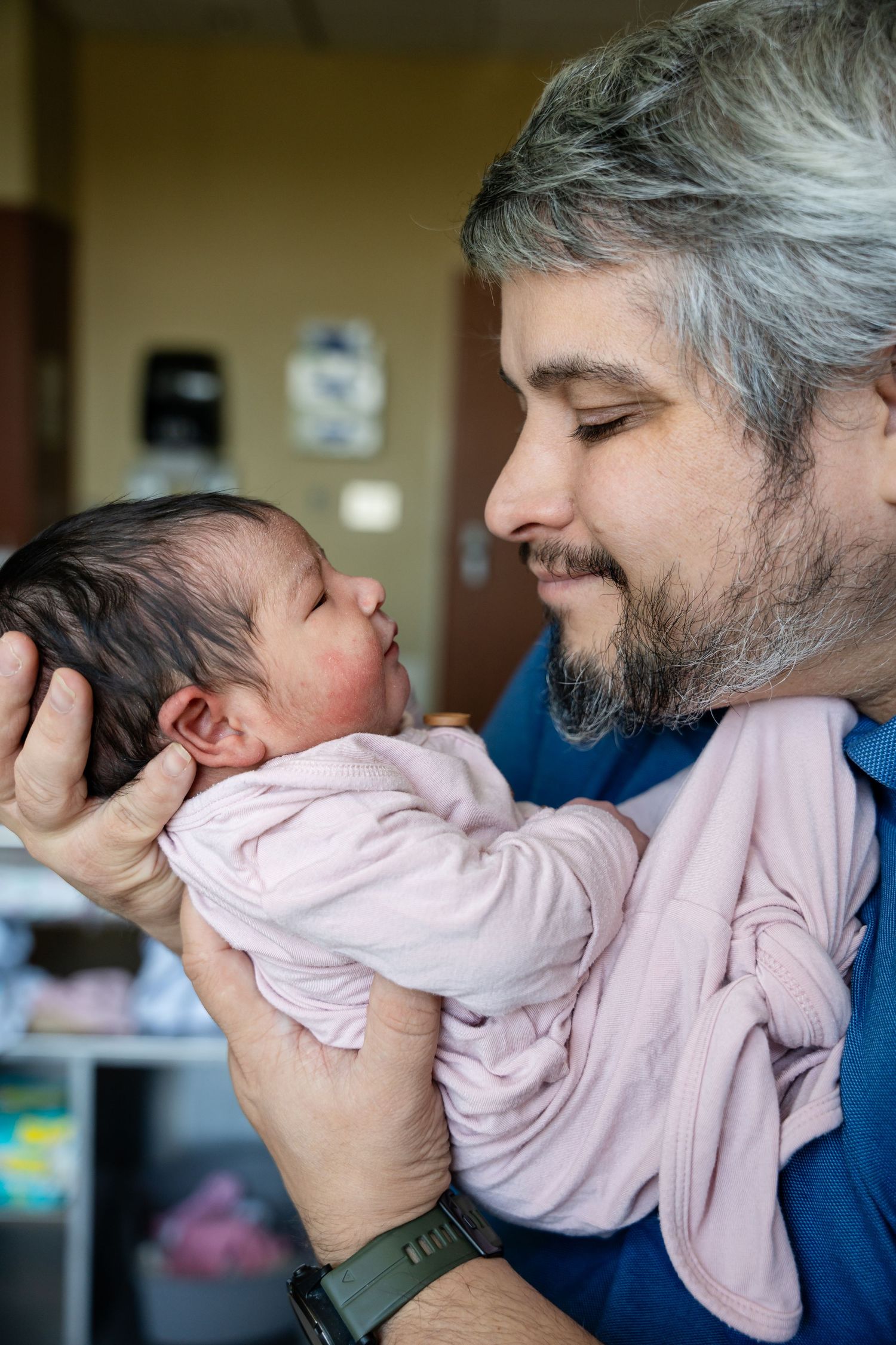 Gray-haired bearded father gently holding a sleeping newborn baby in a pink outfit up to his face in a hospital room.