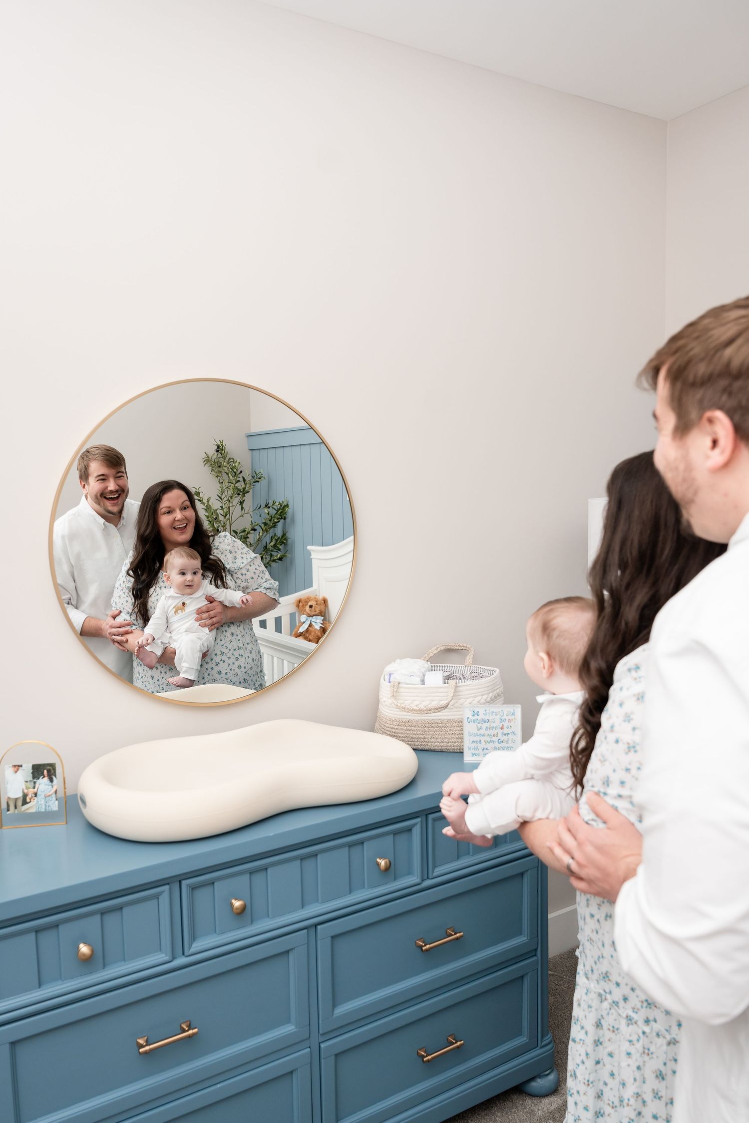 A nursery scene with a blue dresser and round mirror reflecting a family moment.