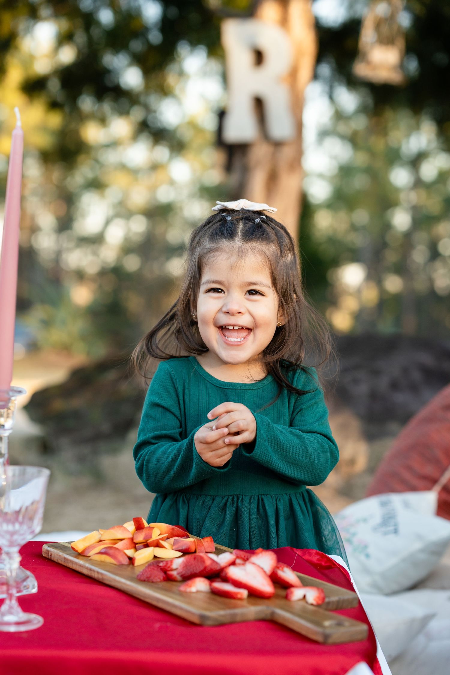 Young child in green dress enjoying food from wooden serving board with red tablecloth at outdoor picnic.