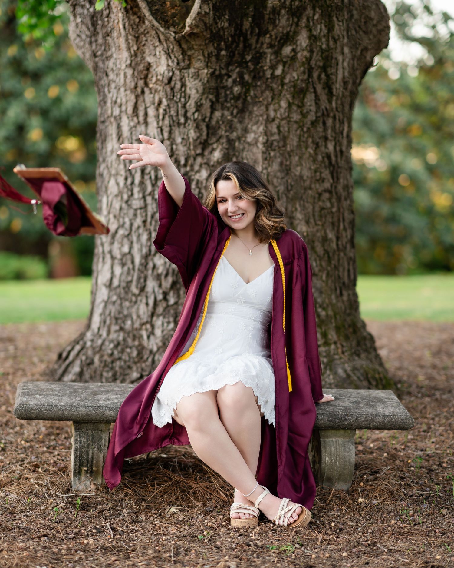 Graduate sits casually by a large tree trunk wearing a burgundy gown and white dress.