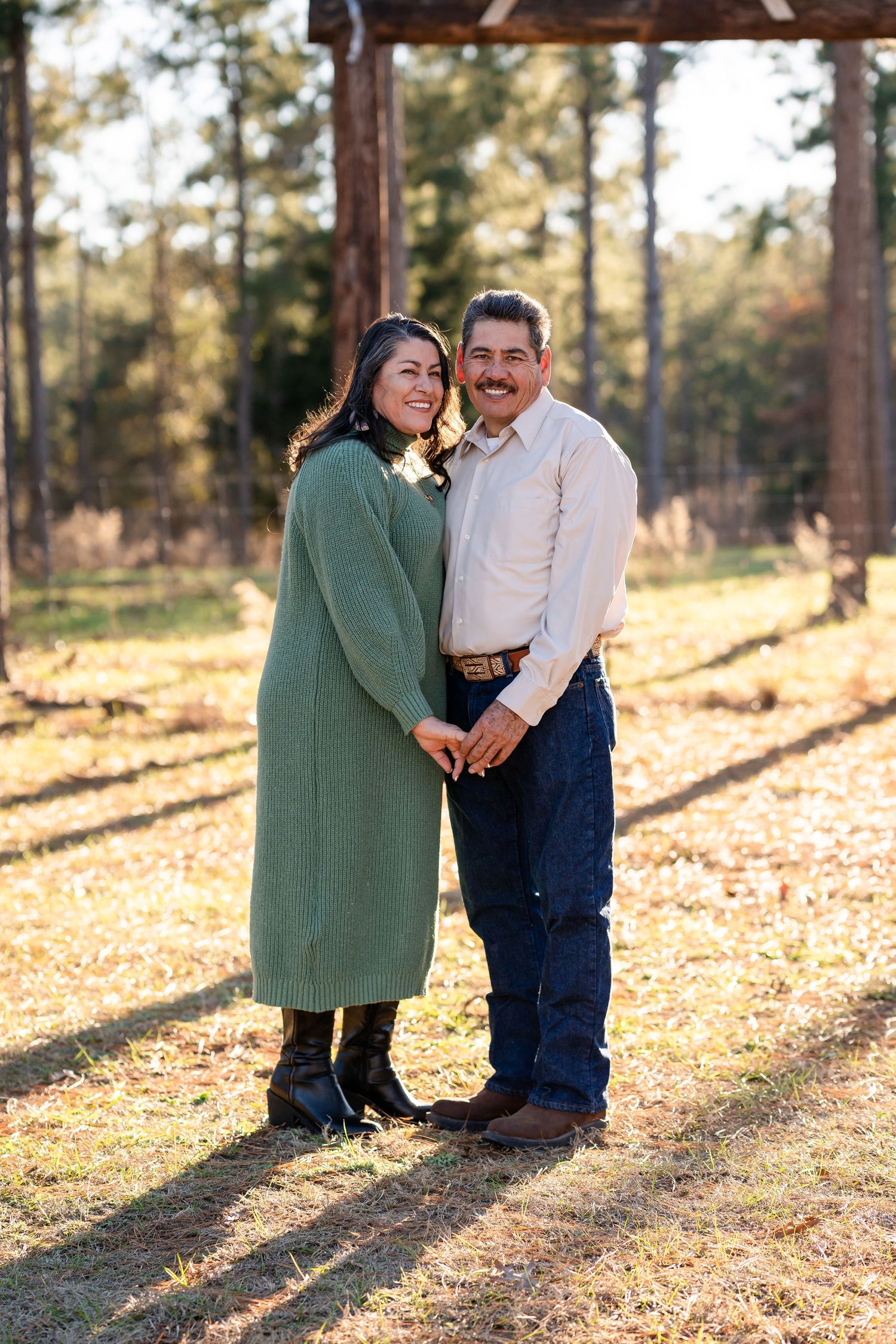 A couple embraces while standing in a wooded area with golden sunlight filtering through the trees.