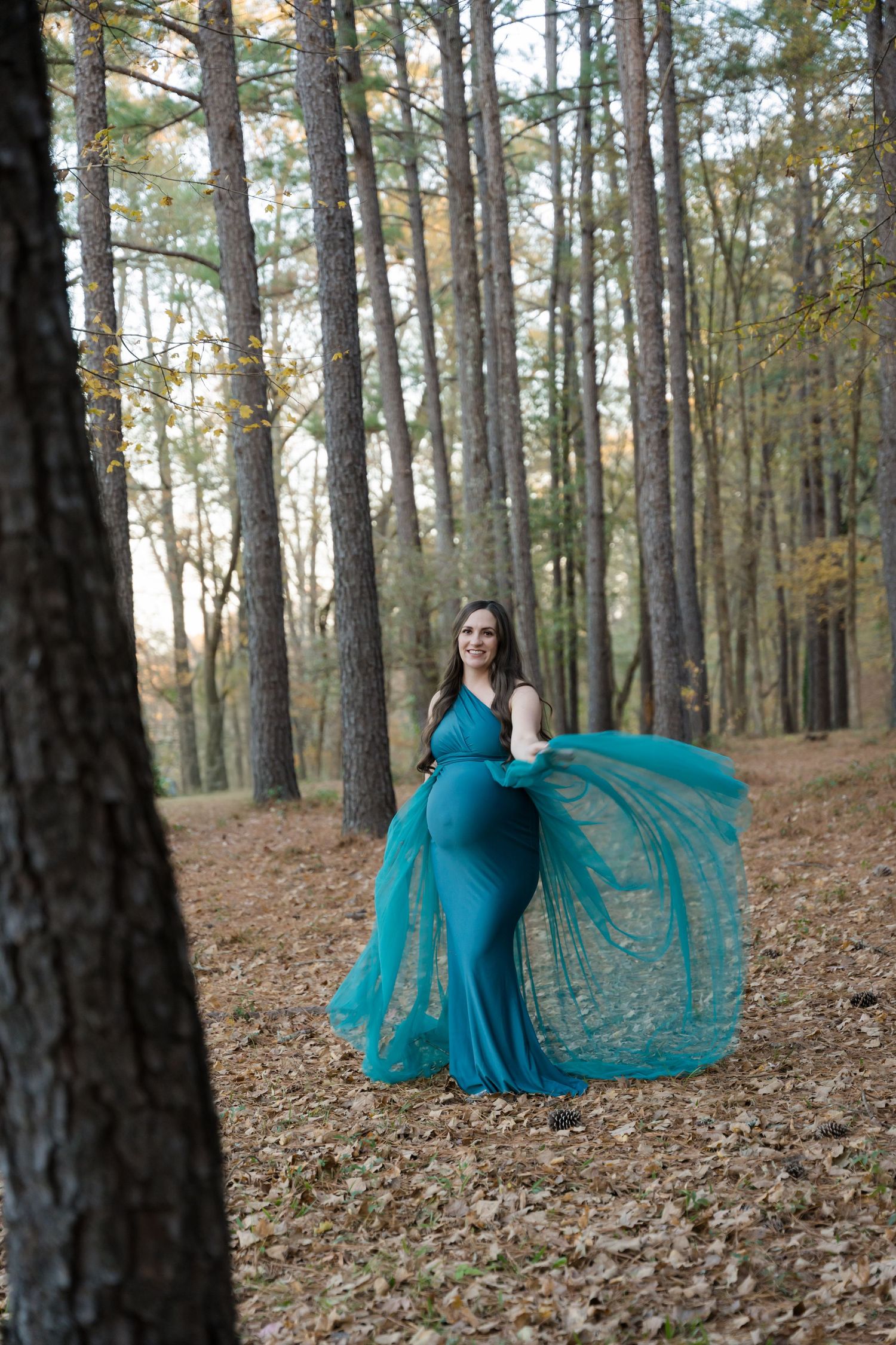 A flowing teal dress creates an ethereal effect against a backdrop of tall pine trees in an autumn forest.