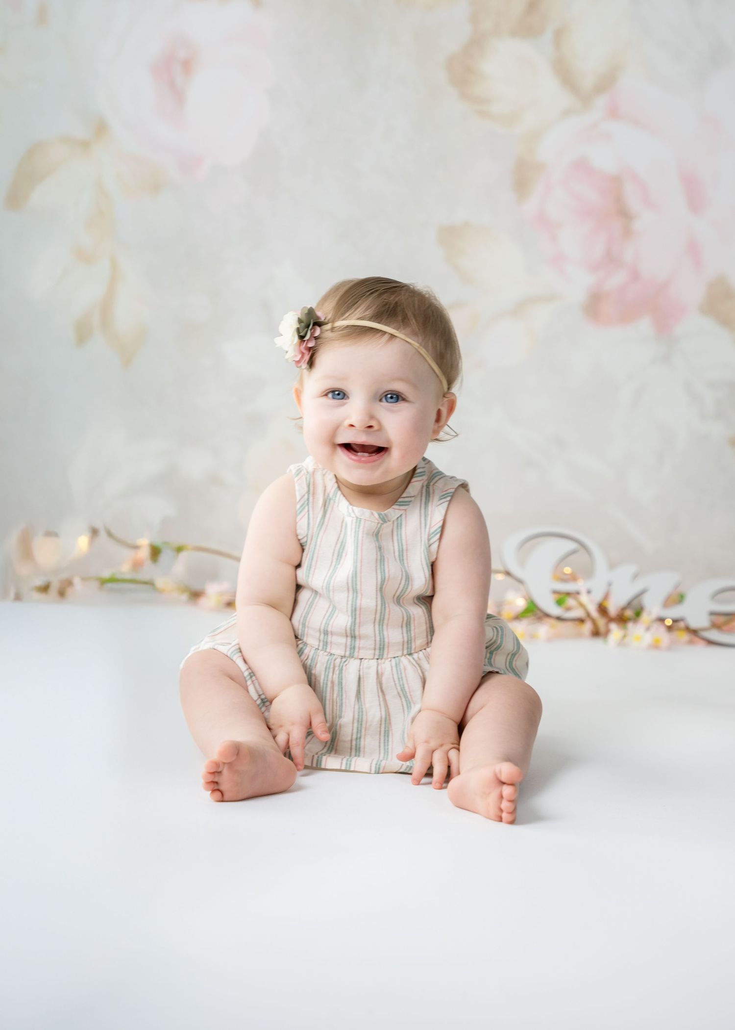 Adorable little one in striped romper sitting on white floor with floral backdrop.