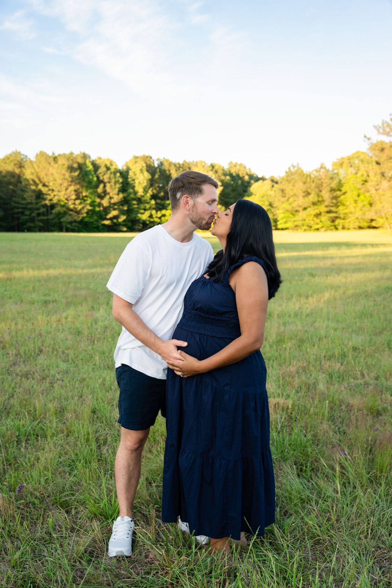 Expecting couple sharing a kiss in a sunny grassy field with trees behind them, the woman in a navy maxi dress cradling her baby bump.