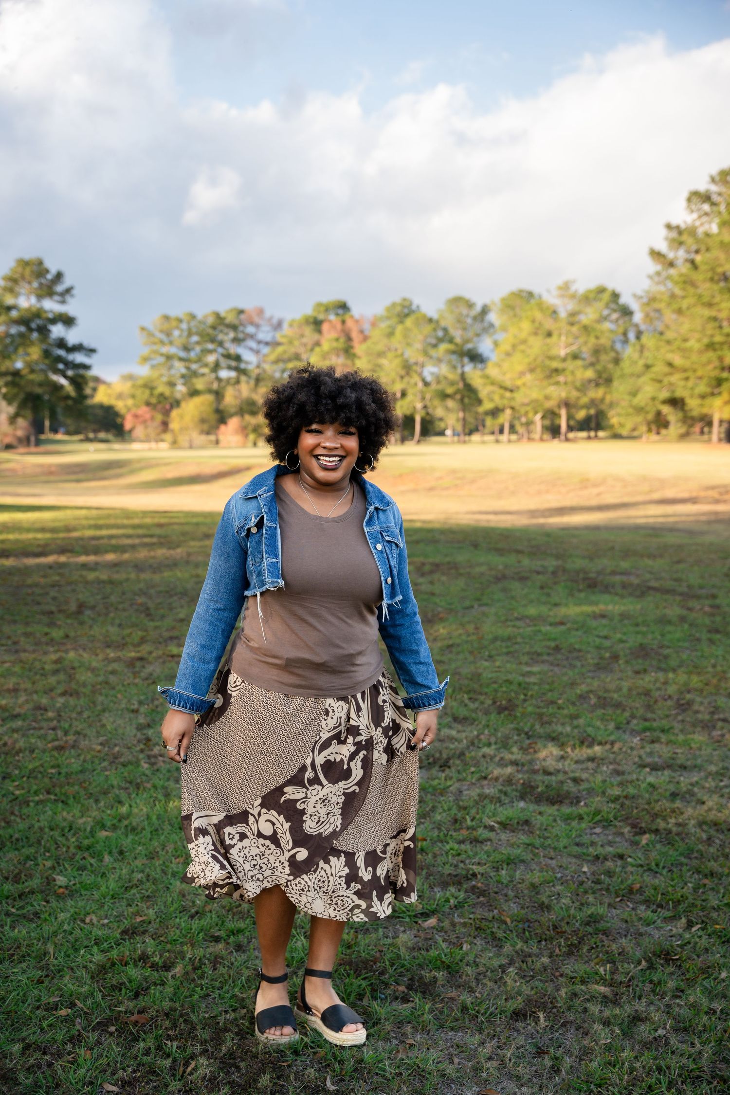Person wearing denim jacket and patterned dress poses playfully in grassy outdoor setting.