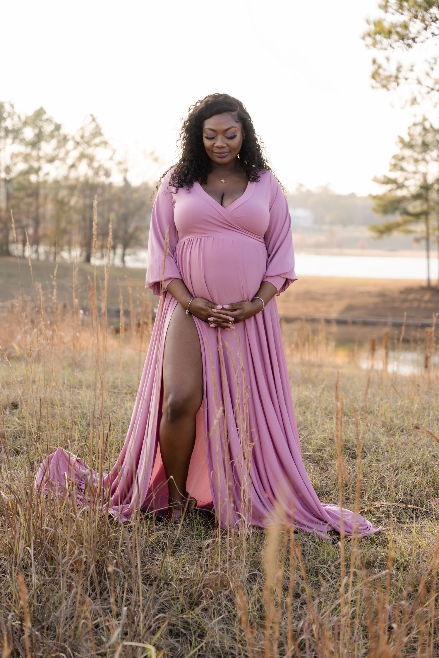 Pregnant woman in flowing pink maternity gown standing in golden field at sunset.