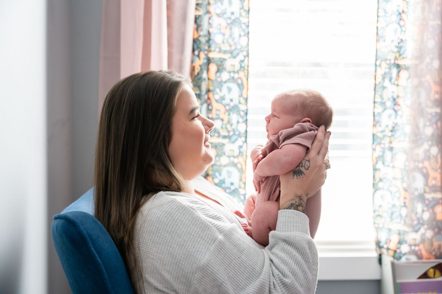 A young mother holds her newborn baby up to her face while sitting by a window with floral curtains.