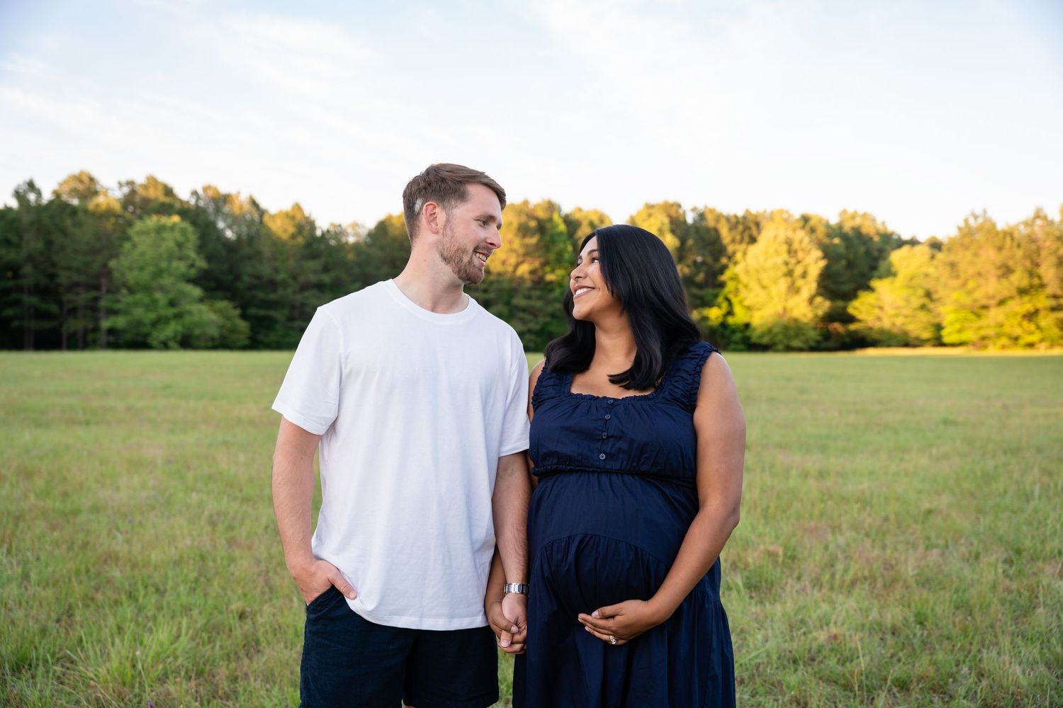 Couple smiling at each other in a sunny field, pregnant woman in navy dress holding her baby bump.