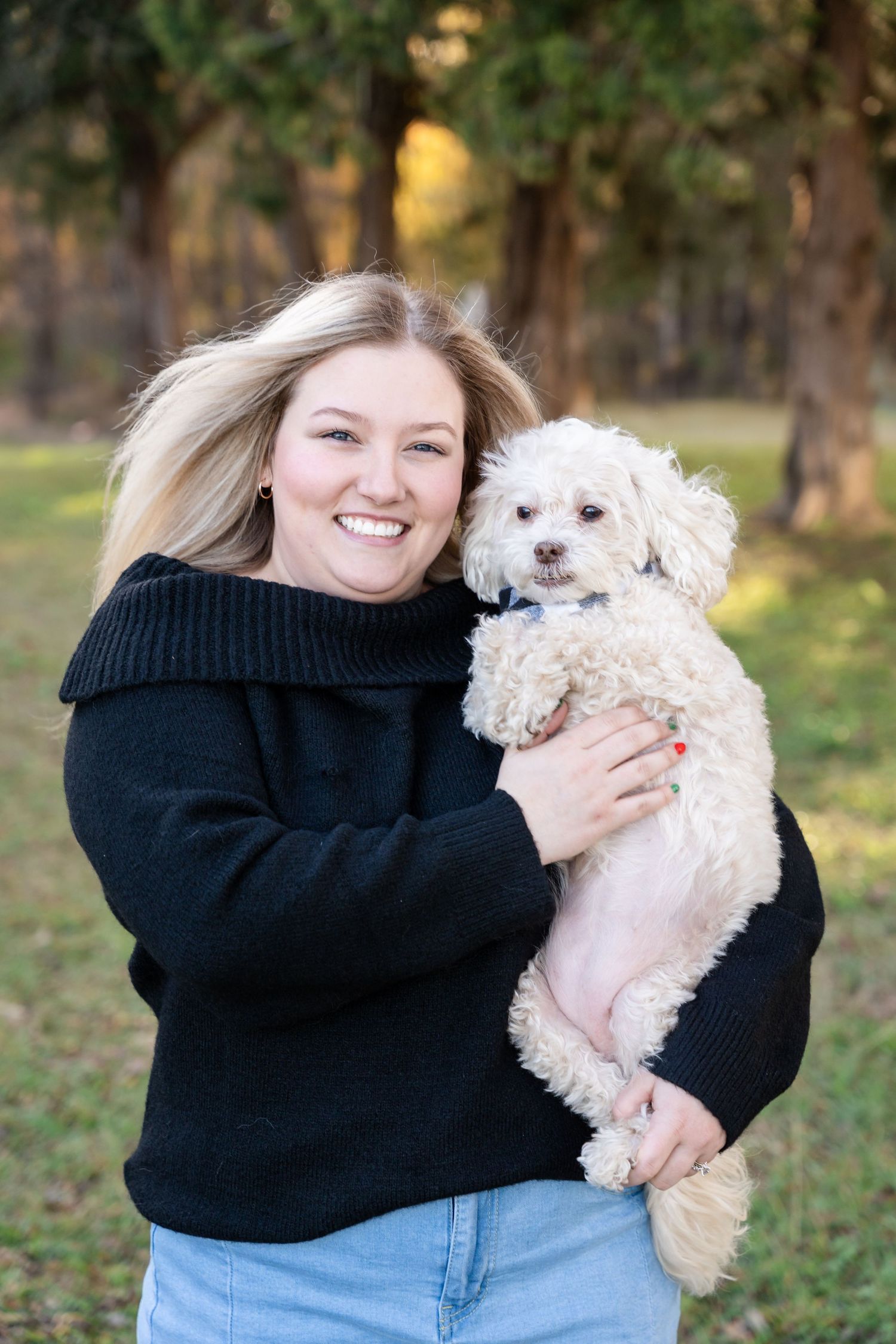 Smiling woman with long blonde hair in a black sweater holds a fluffy white dog outdoors on a grassy lawn surrounded by trees.
