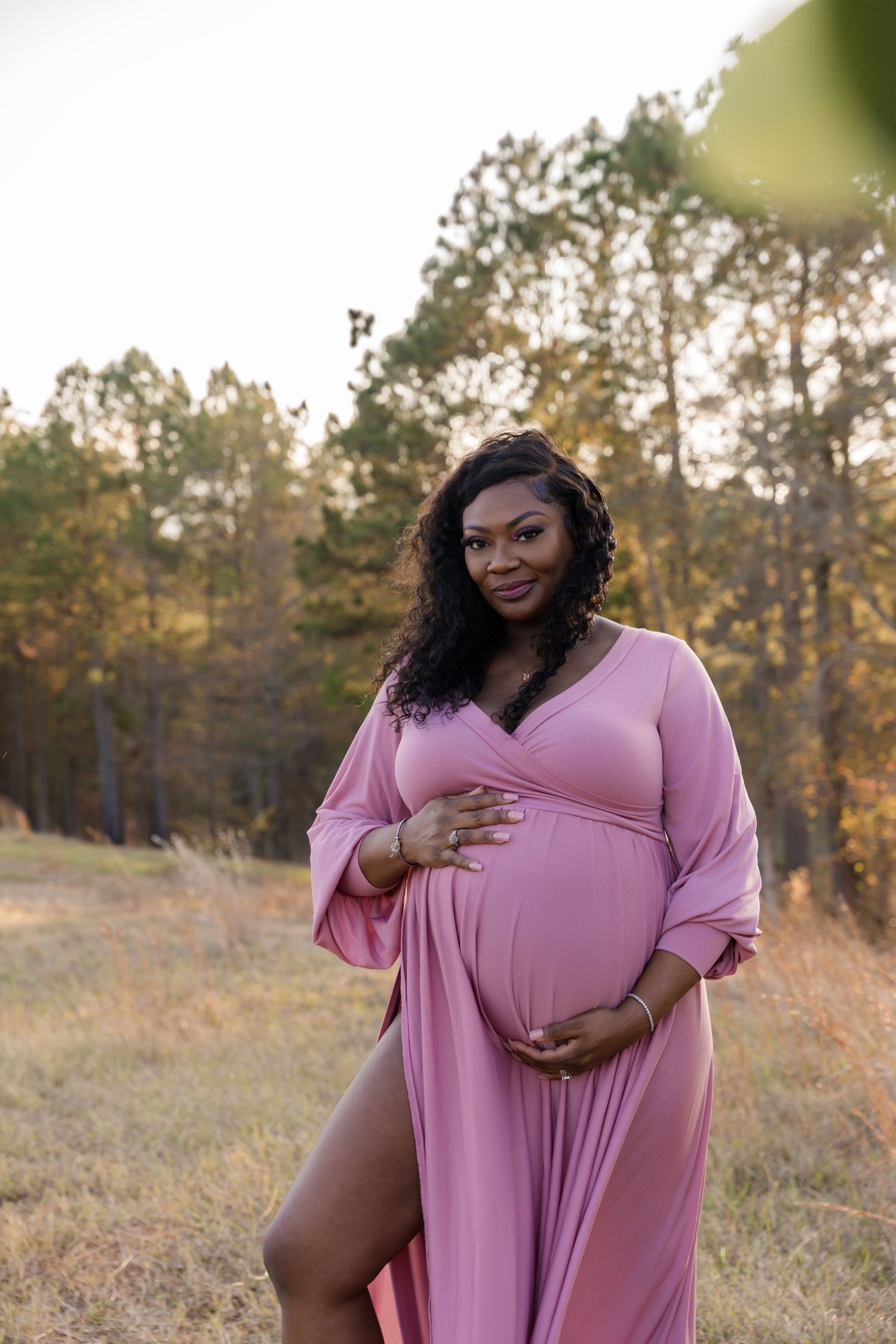 Pregnant woman in pink maxi dress holding baby bump outdoors in autumn field with trees.