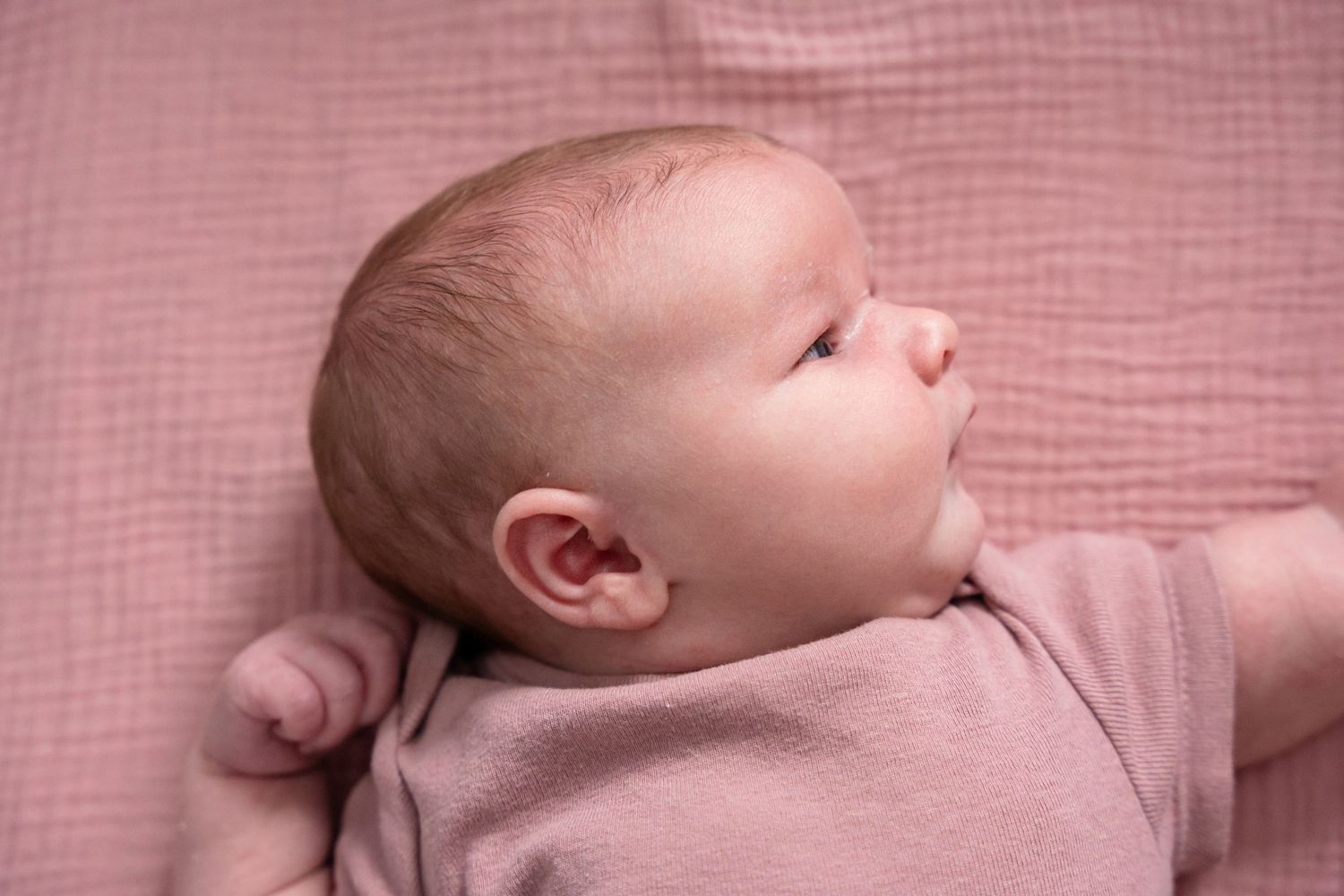 Newborn baby lying on a pink blanket wearing a pink onesie, looking upward with wide eyes.