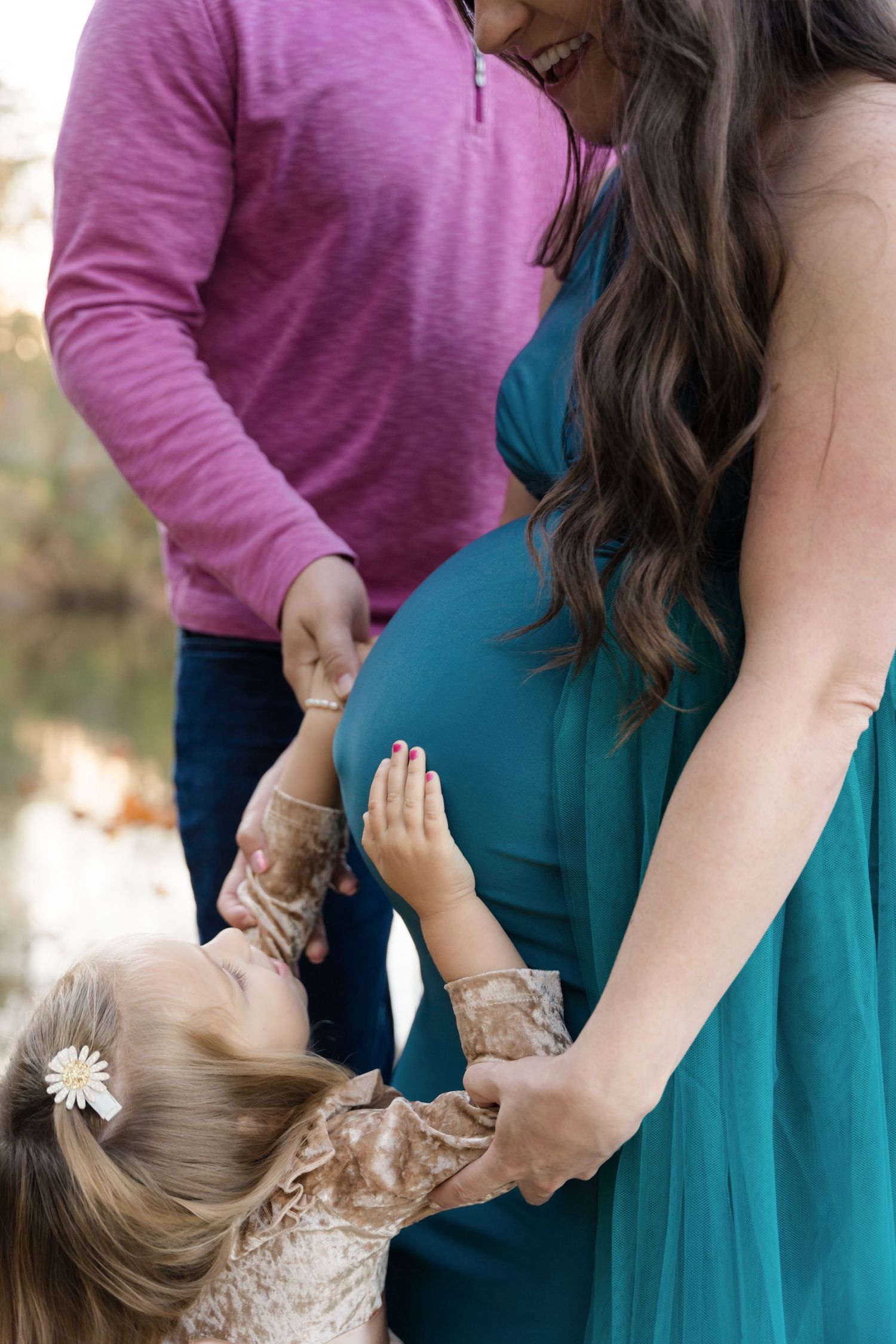 A small child in a lace dress touches the belly of a woman wearing a teal maternity dress while someone in pink looks on.