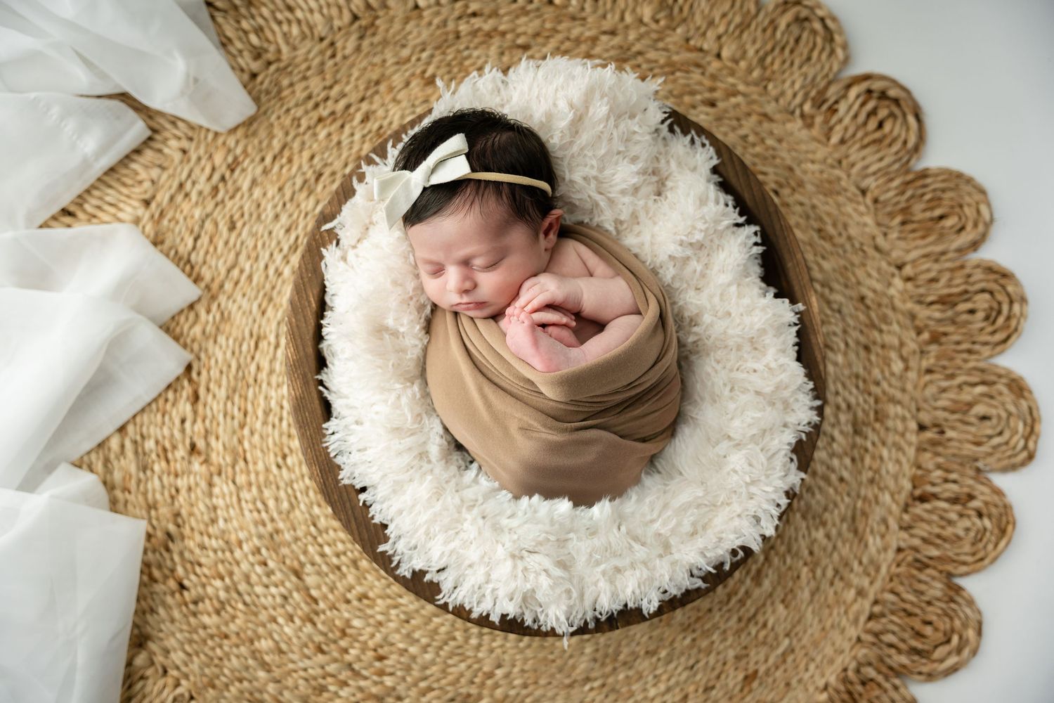 Newborn baby sleeping peacefully on white fluffy blanket with tan wrap and headband on round woven mat.