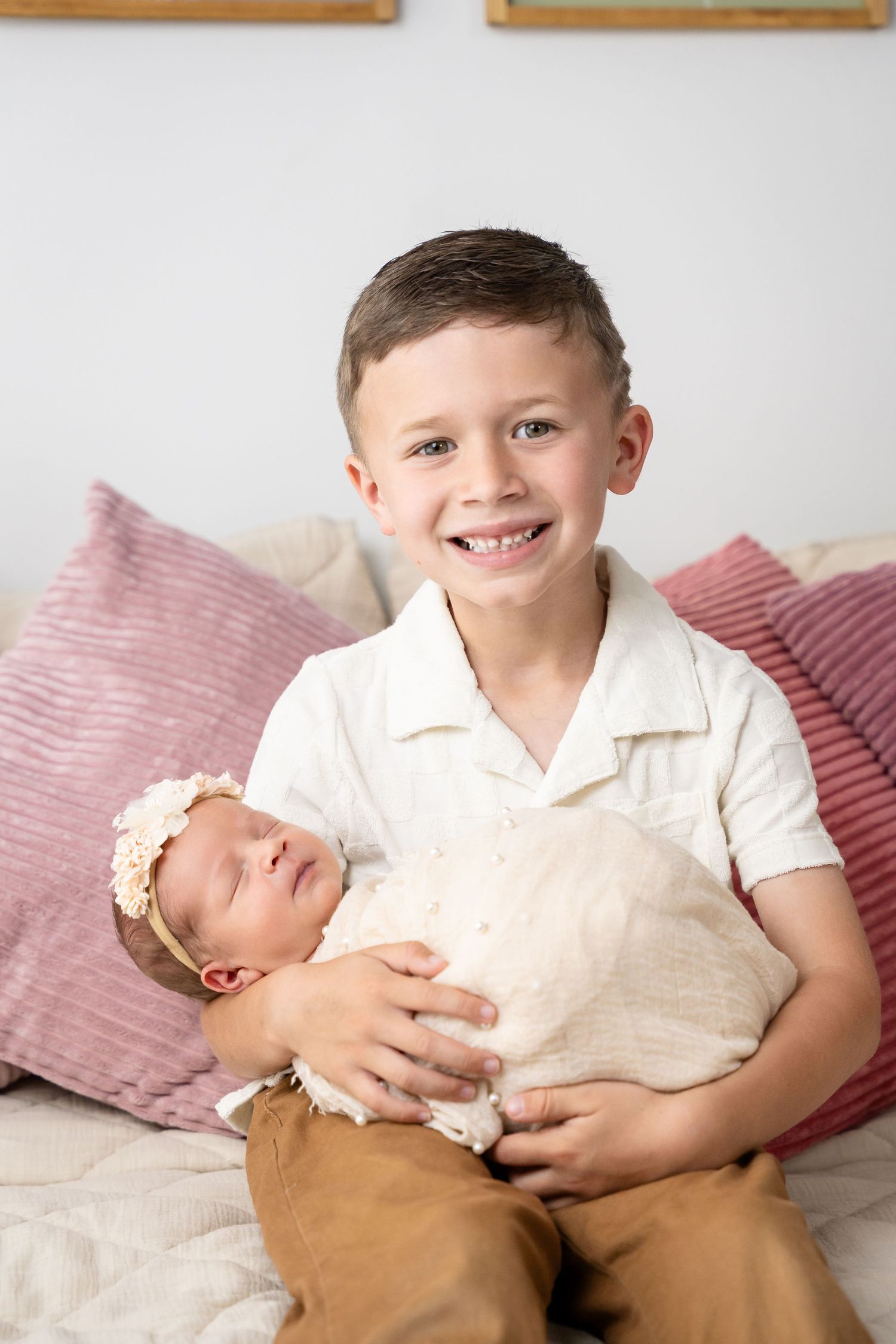 Child in white shirt and brown pants holding newborn sibling against pink cushions.
