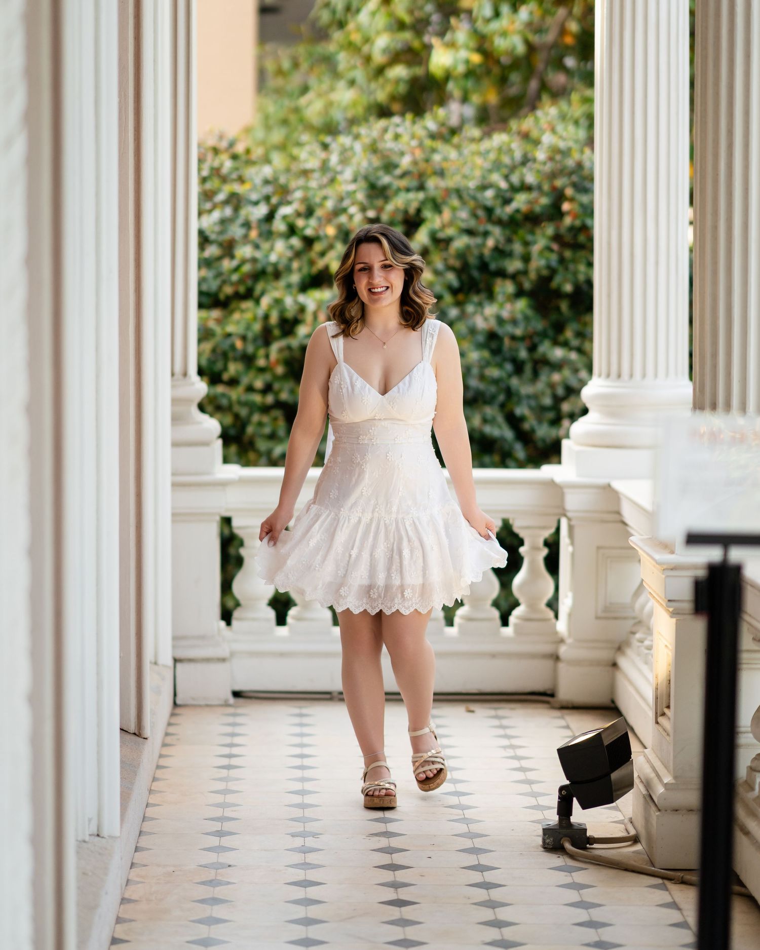 Graduate walks gracefully through a classical colonnade with white pillars and tiled floor.