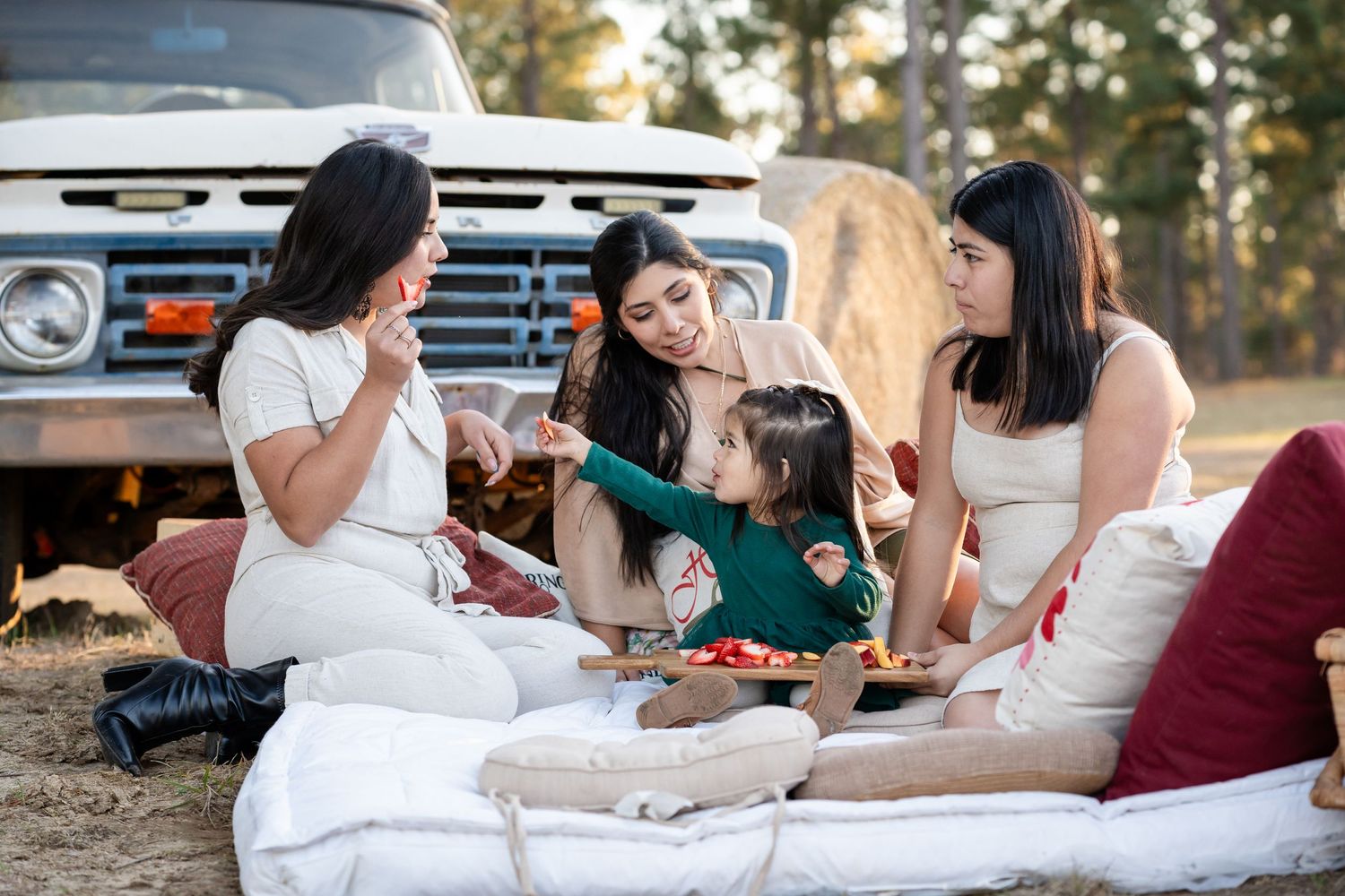Group enjoying outdoor picnic on white blankets near vehicle in sunlit forest setting.