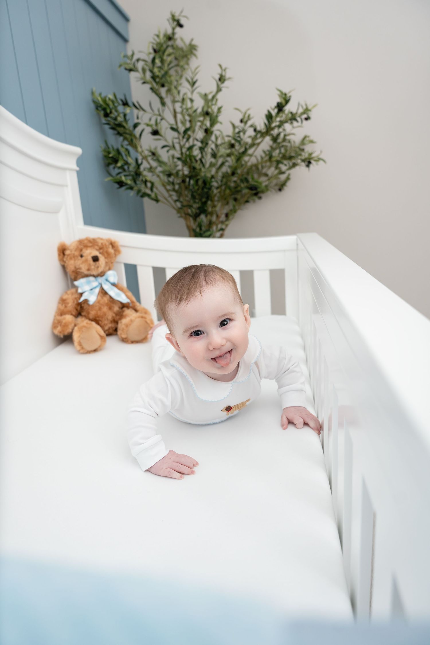 Happy baby in white onesie standing in crib with teddy bear and greenery decor.
