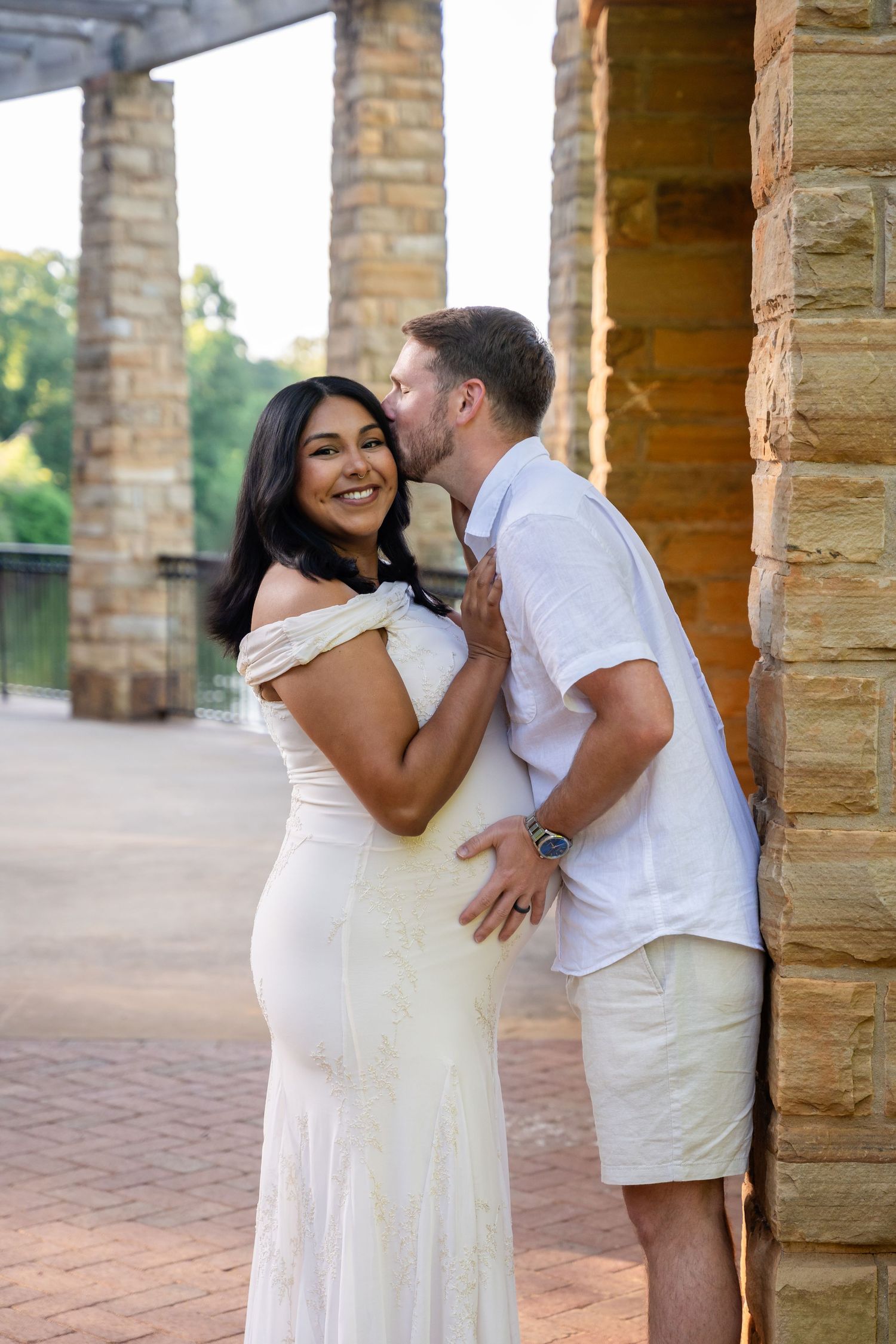 A couple shares a romantic moment by stone pillars, woman in white dress smiling as man whispers in her ear.