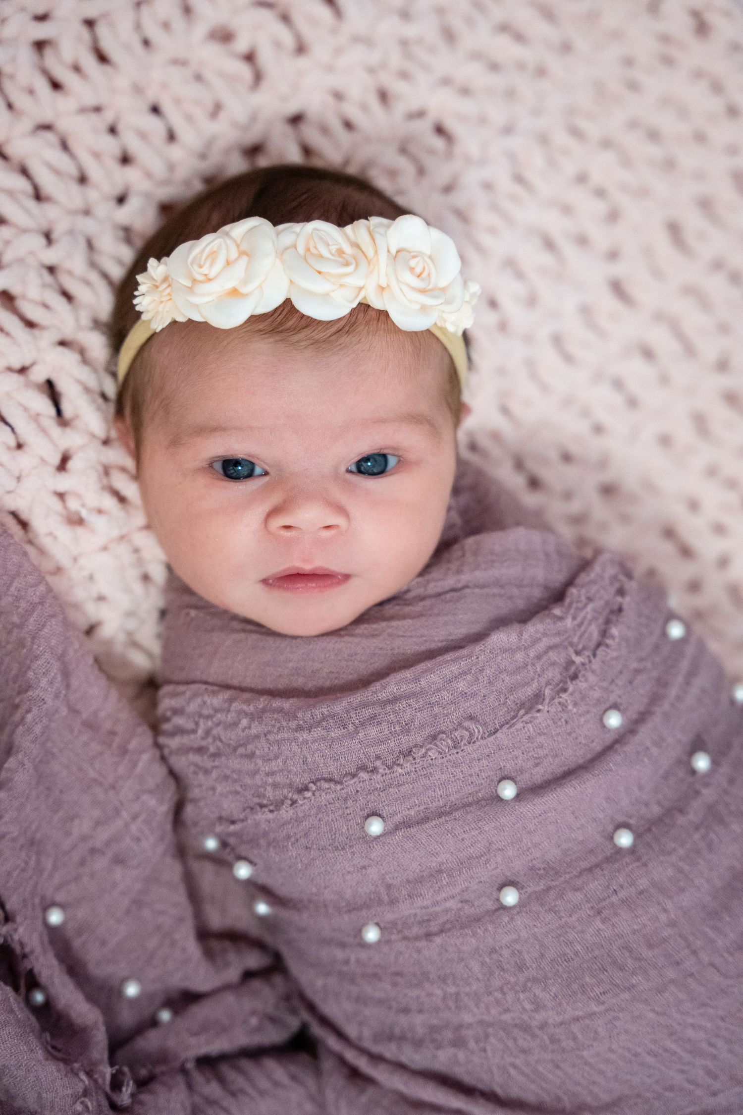 Newborn baby girl swaddled in purple wrap with pearl details, wearing a floral rose headband, lying on pink knit blanket.